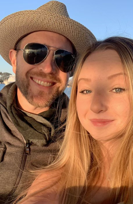 Kennedy Stonum poses for a selfie with her father, Lee, on a sunny day by the water. Lee, standing next to Kennedy on the left, is wearing sunglasses and a straw sun hat.