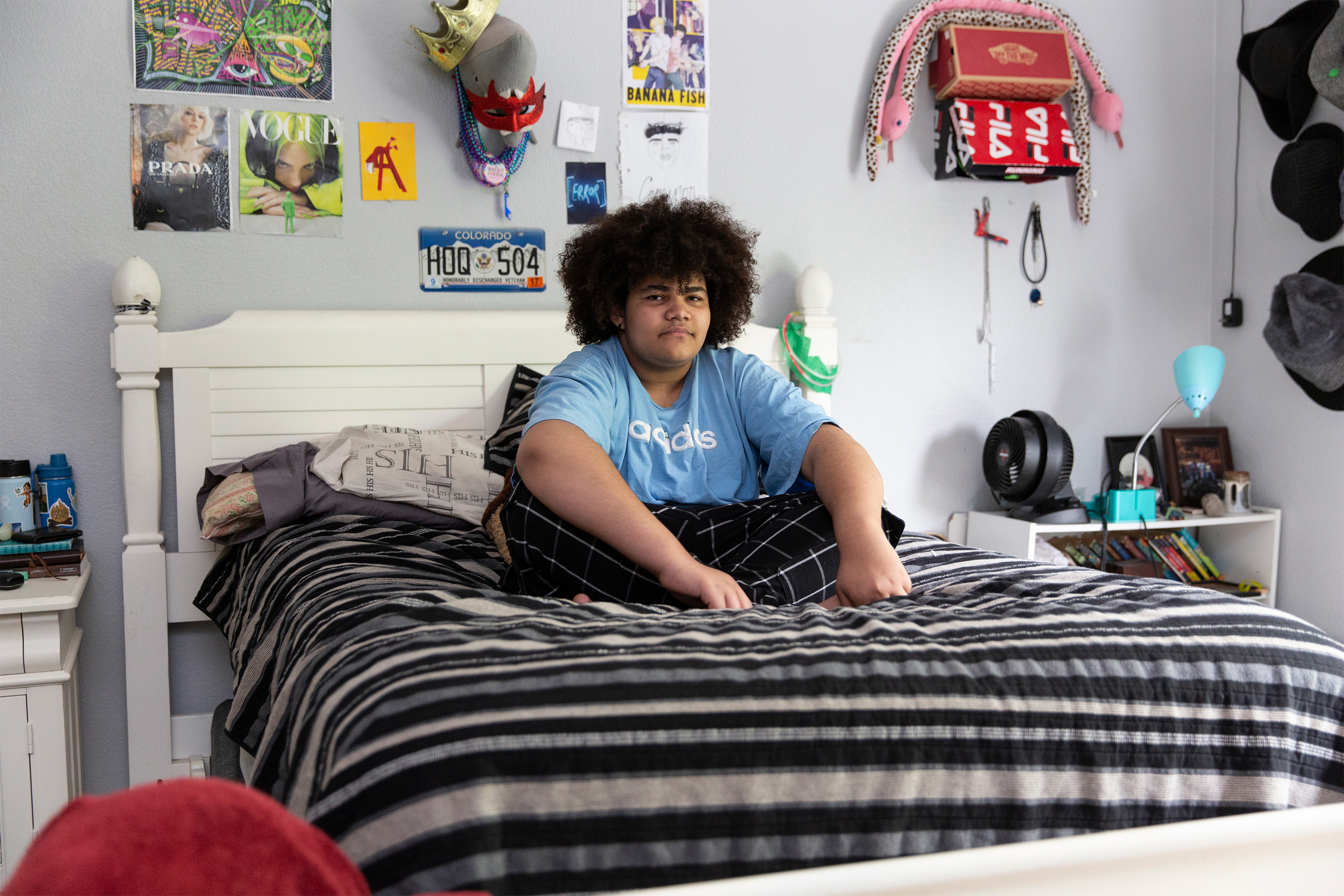 A wide shot photo shows Cameron Wright sitting in his bed at home in Denton, Texas. Posters and trinkets hang on the wall behind him.