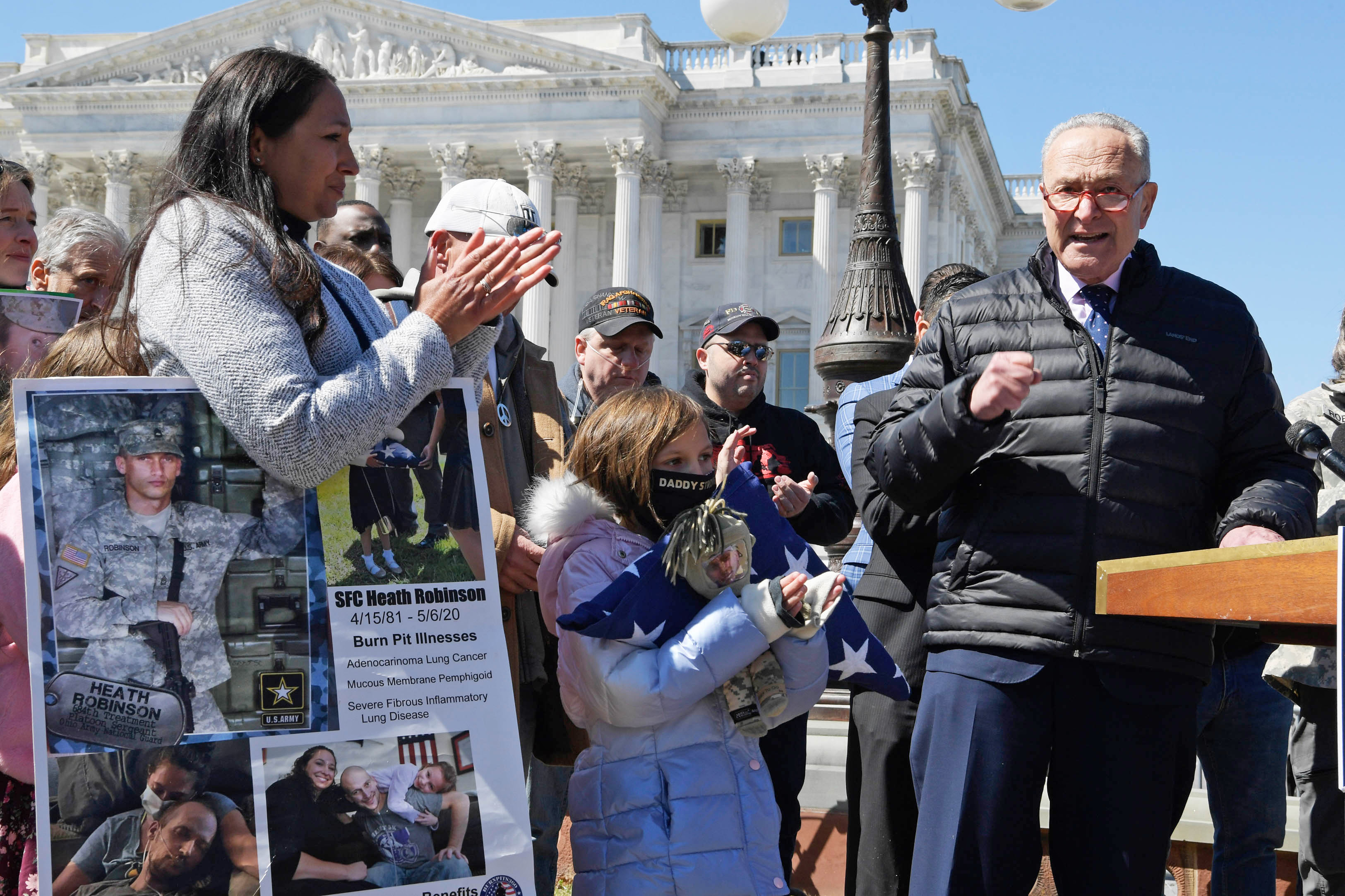 Senator Chuck Schumer is seen standing at a podium with a microphone, talking to a crowd at a press conference. People hold signs and are seen wearing veteran's hats around him.