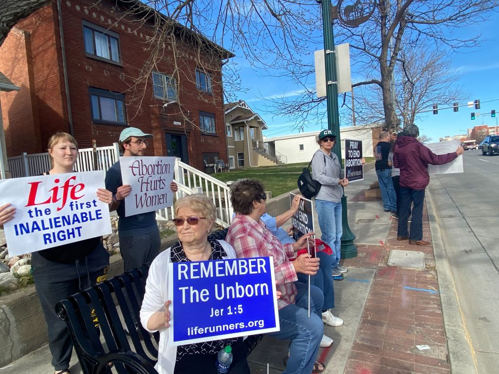 A group of anti-abortion protesters outside of the clinic. They hold signs that read, "Abortion Hurts Women," "Remember the Unborn," "Pray to End Abortion," among others.