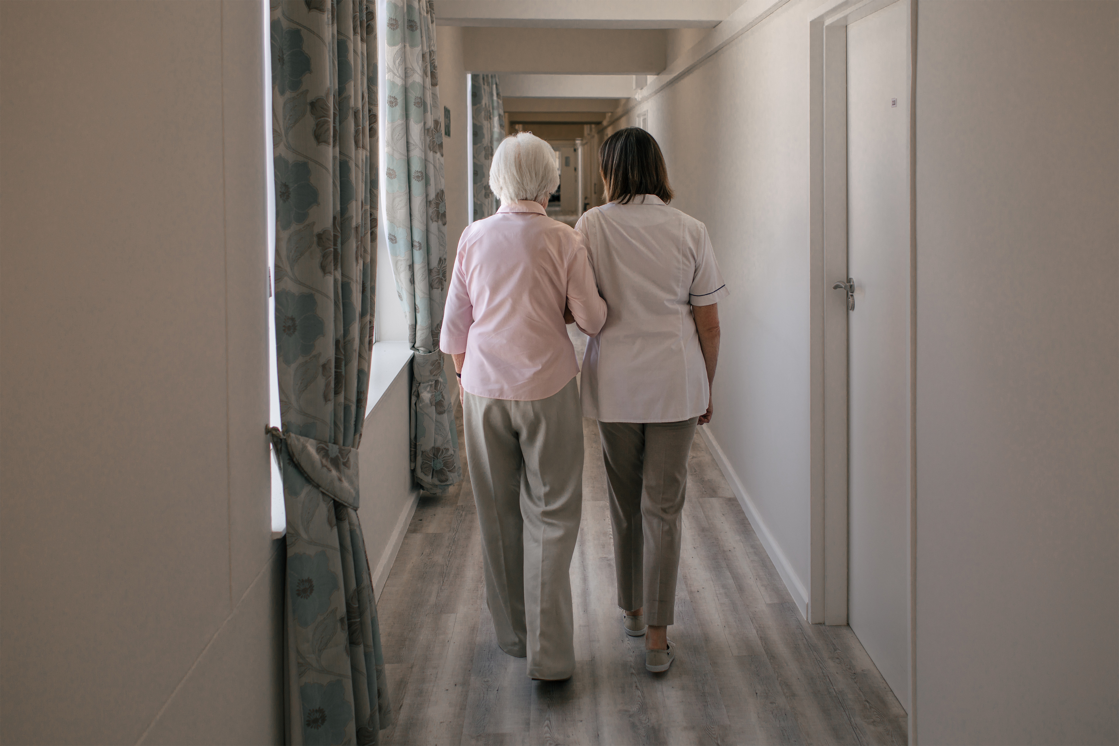 A photo shows a nurse walking an elderly woman down a hallway in a nursing home. The two are seen from behind.