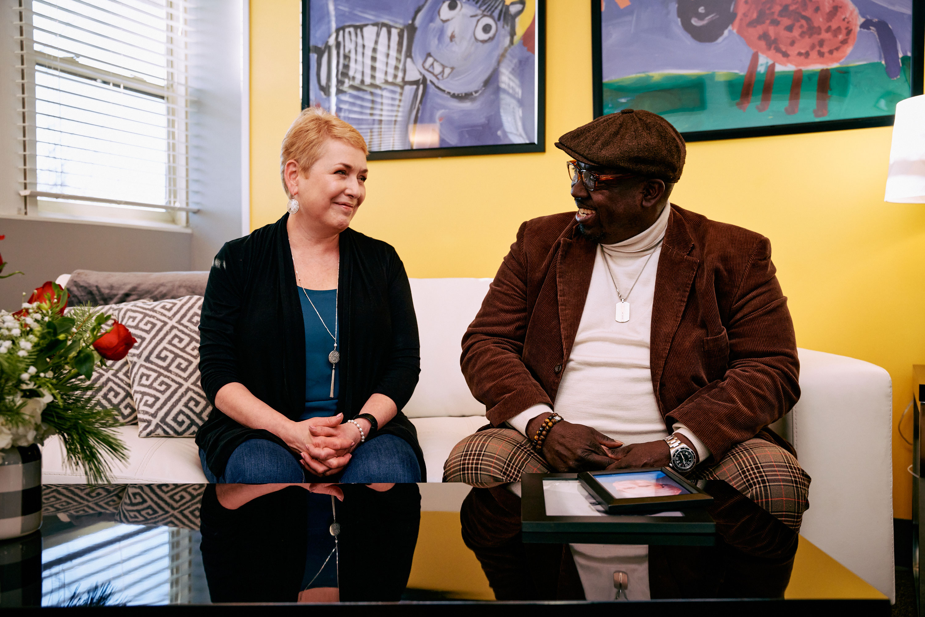 A woman and a man talk while sitting on a white couch in a colorful room.