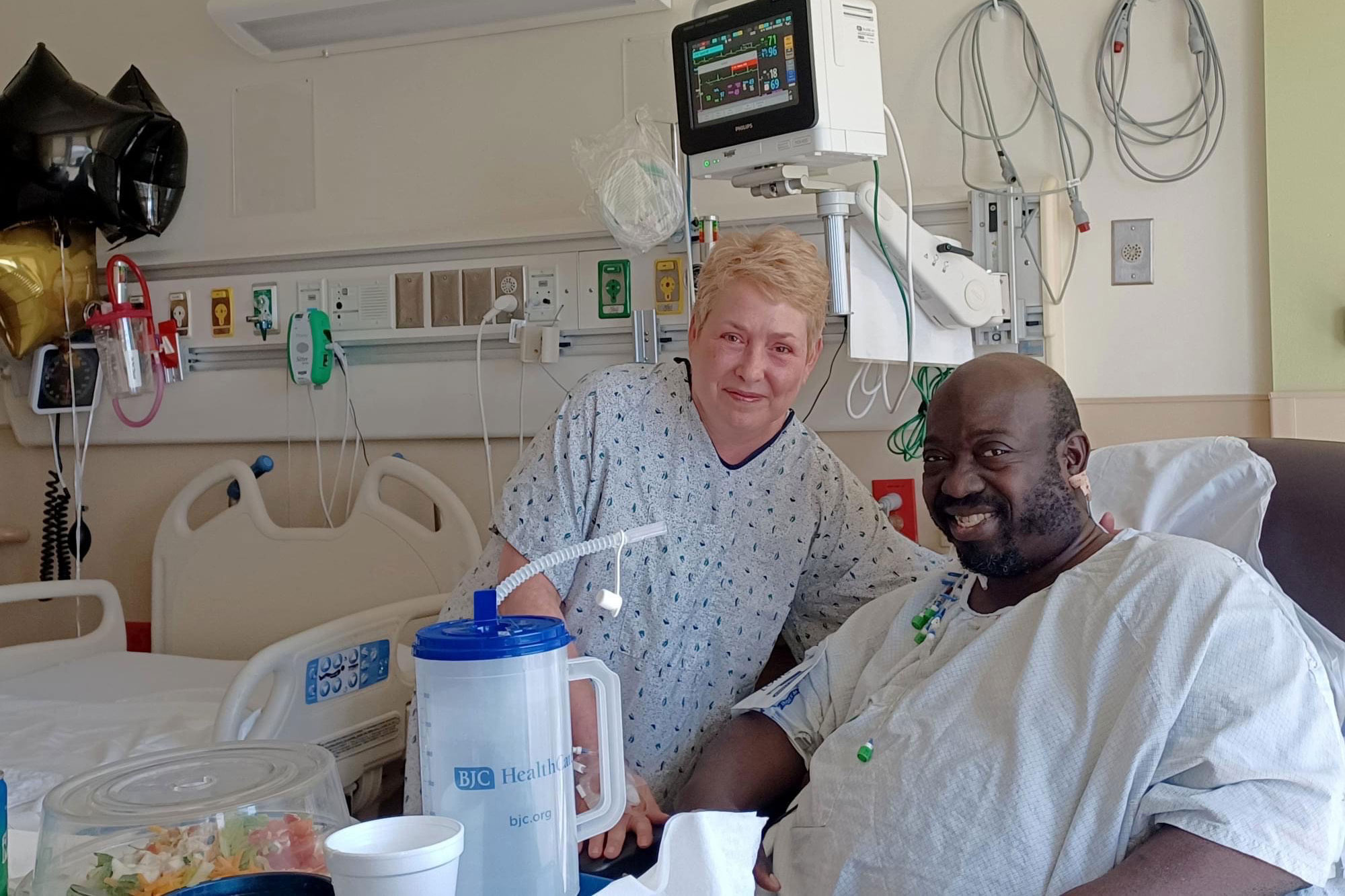 A woman and man in hospital gowns in a hospital room pose together.
