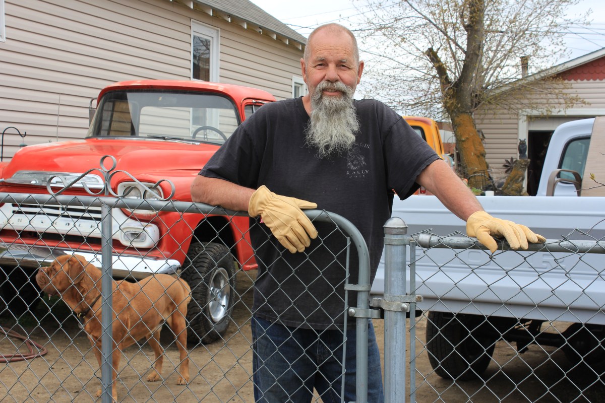 Al Shields stands in the center of the photograph behind a chain-link fence, which he rests his hands on. Behind him are two vintage trucks, one a soft red and the other pale blue.