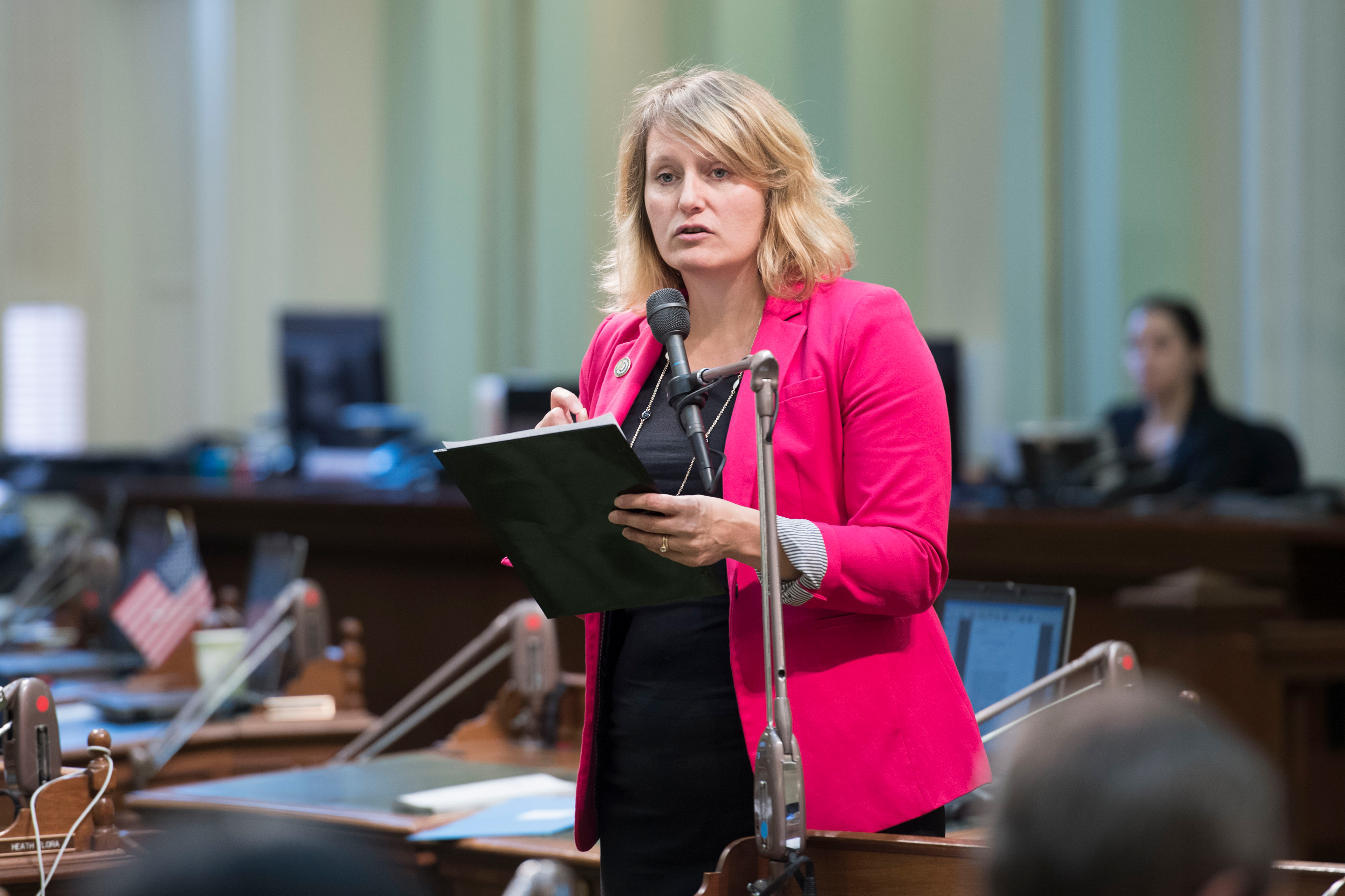 Buffy Wicks is seen speaking in front of a microphone before the California Assembly.