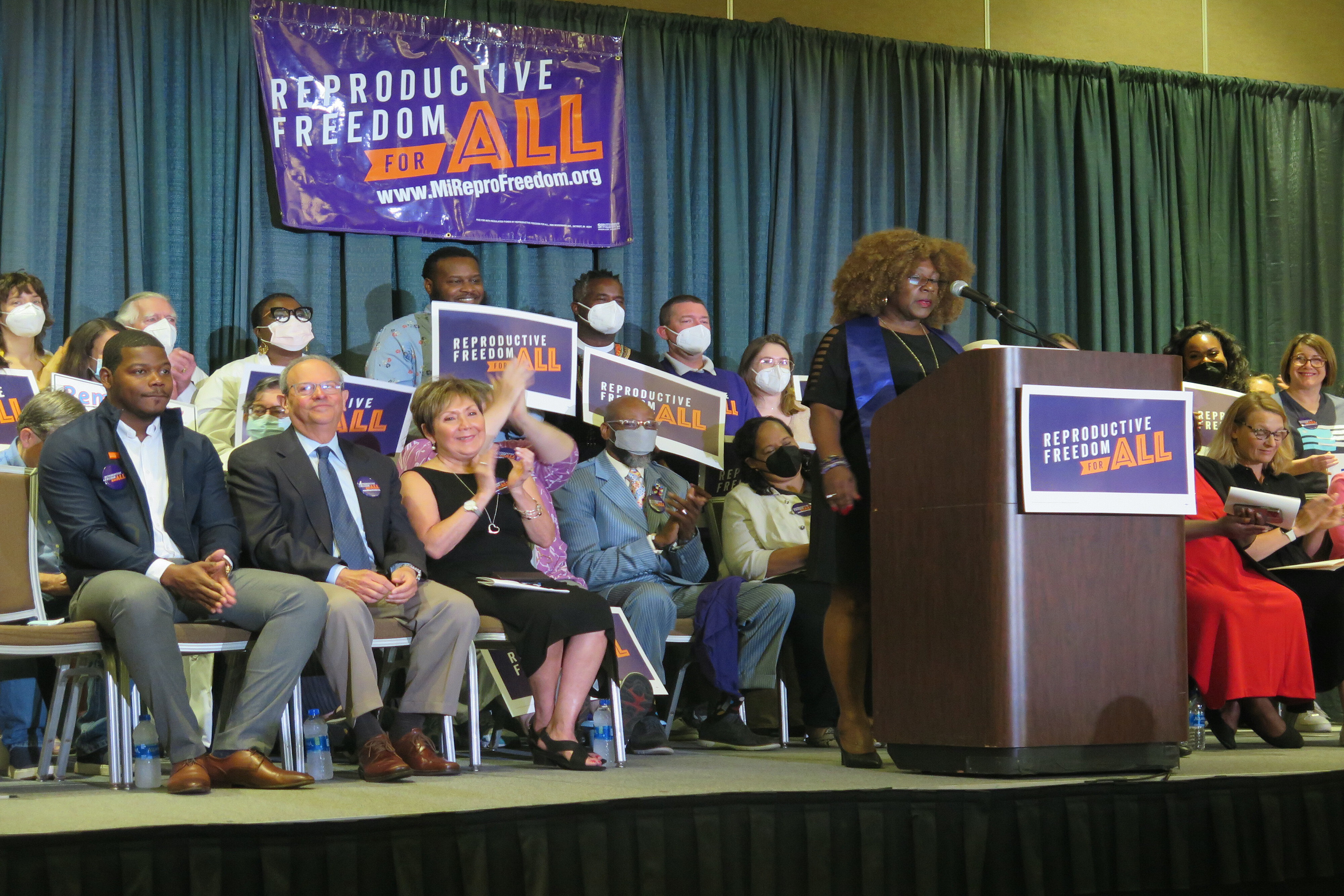 Abortion activist Leslie Mathews stands at a podium in front of a sign that reads "Reproductive Freedom For All". A group of about eight people, holding similar reading signs, are seated on the stage behind here.