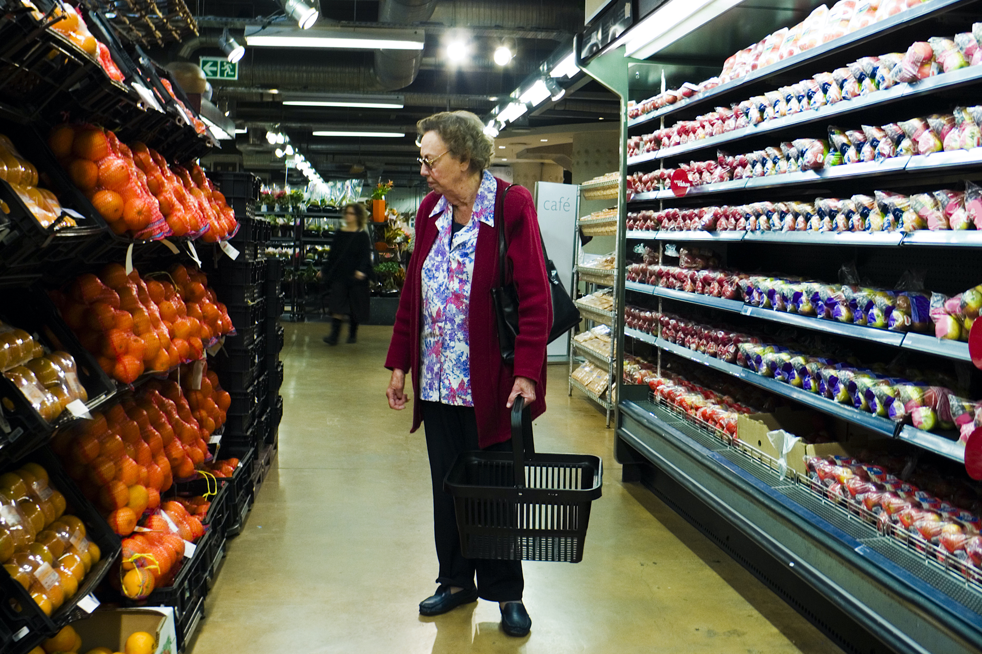 A senior woman stands in a grocery aisle holding an empty shopping basket.