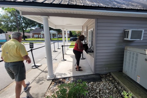 a man in a yellow shirt walks toward a white wood building where a woman in a red tank top is standing at a window speaking to someone inside the building