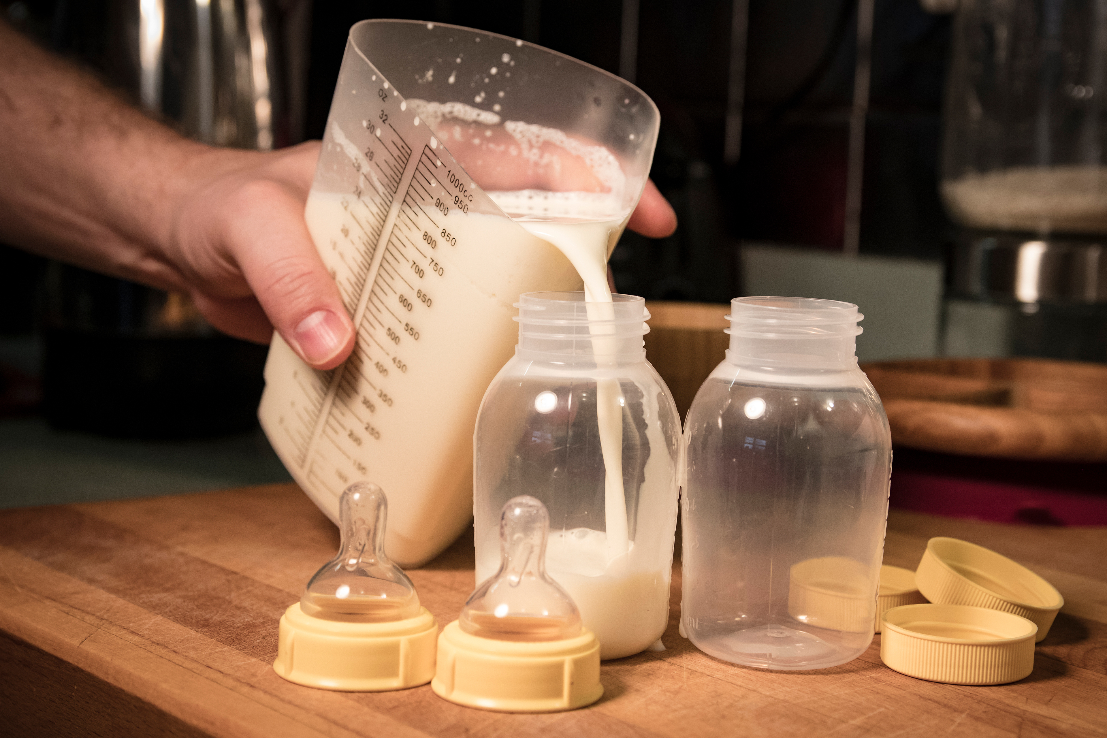 Nutritional formula is poured into a baby bottle. There is a second, empty bottle beside the one being filled. They sit on a wooden countertop.