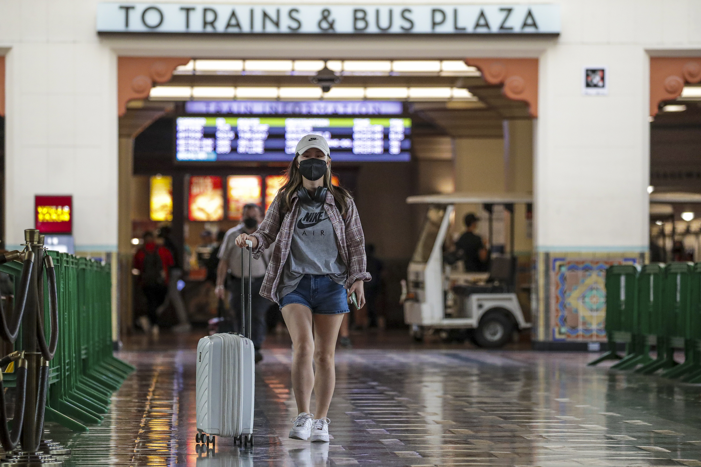 A woman a black face mask rolls a suitcase beside her. A sign behind her reads "To trains & bus plaza".