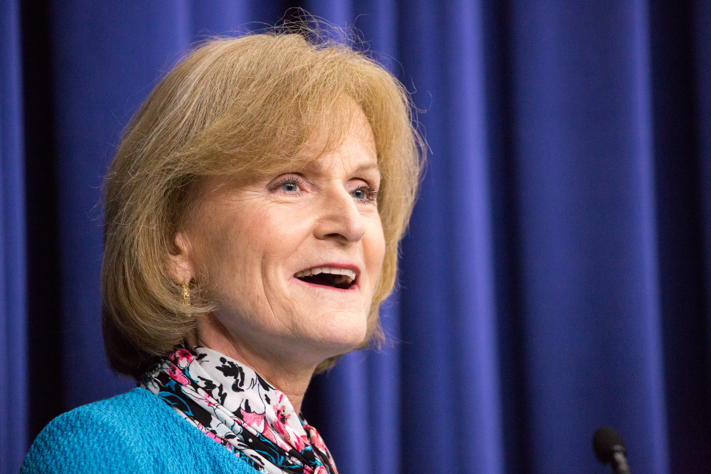 A photo shows Mary Wakefield speaking at a podium with a blue curtain backdrop behind her.