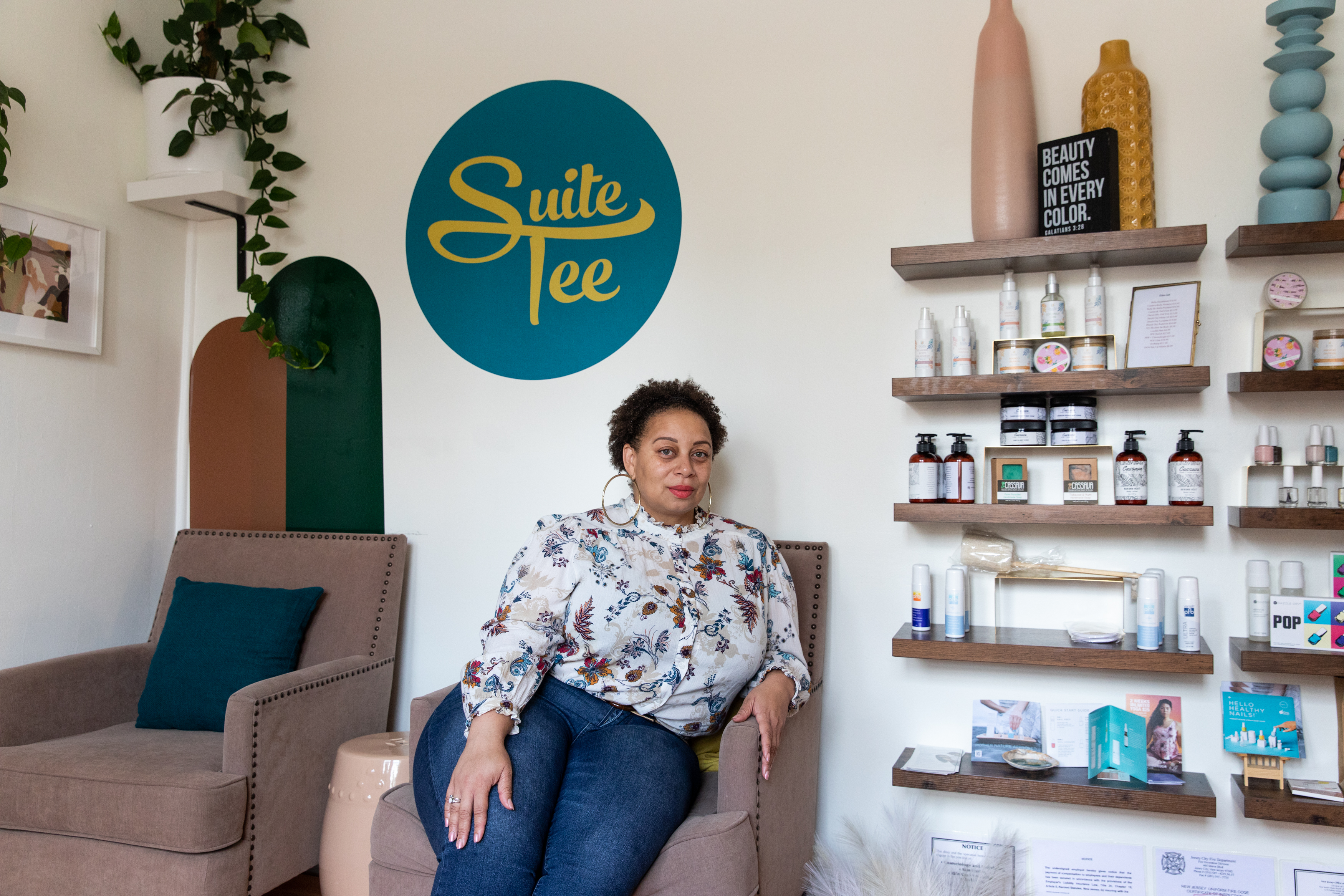 Tee Hundley, an adult female, sits in a chair in her nail salon. She has short, curly hair, wears large hoop earrings, a white blouse with a floral pattern, and jeans. The wall beside her holds nail products, and behind her is a sign that says, "Suite Tee".