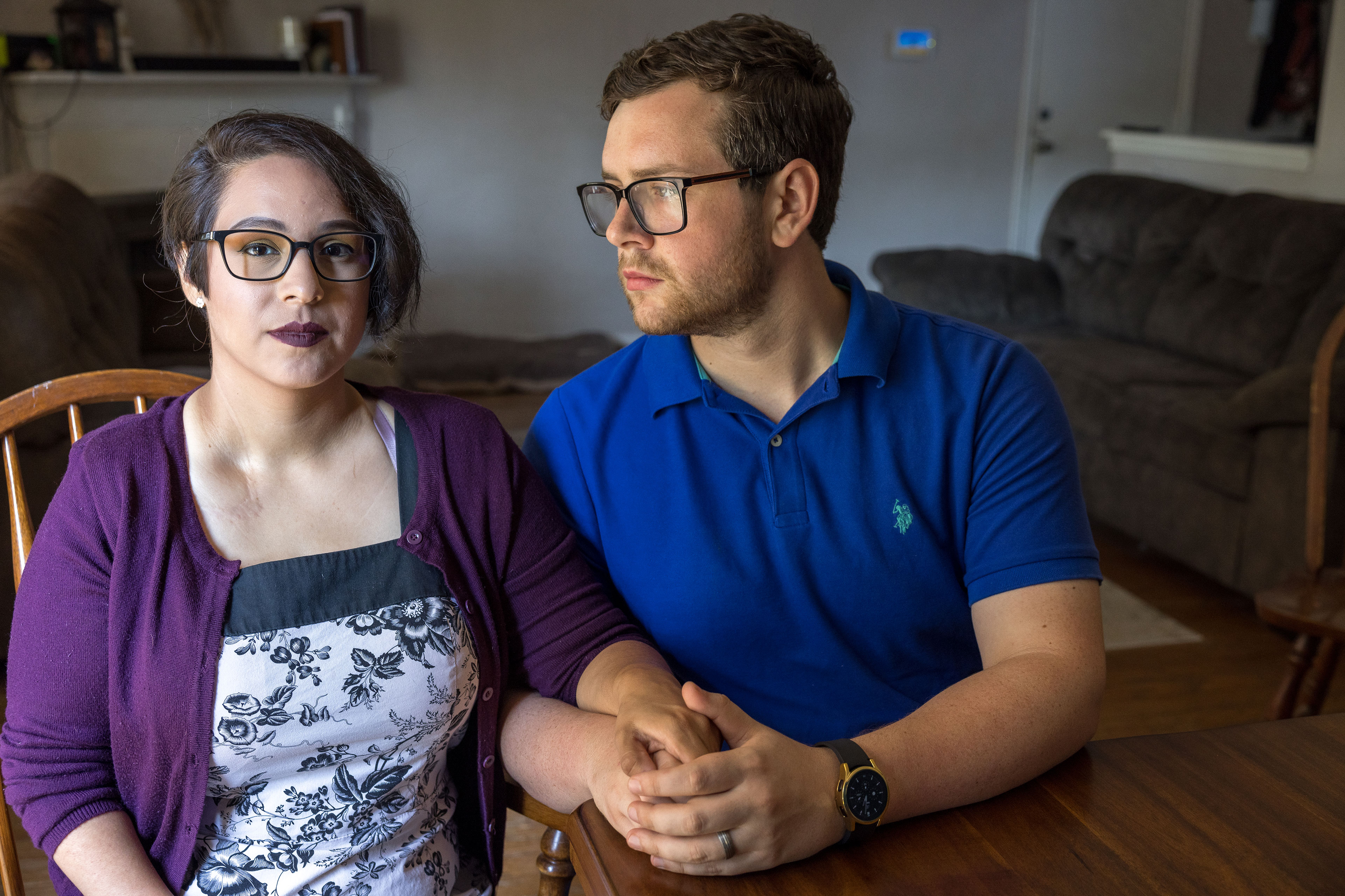 A woman and a man sit at a dining table. The man holds one of her hands in both of his and looks at her.