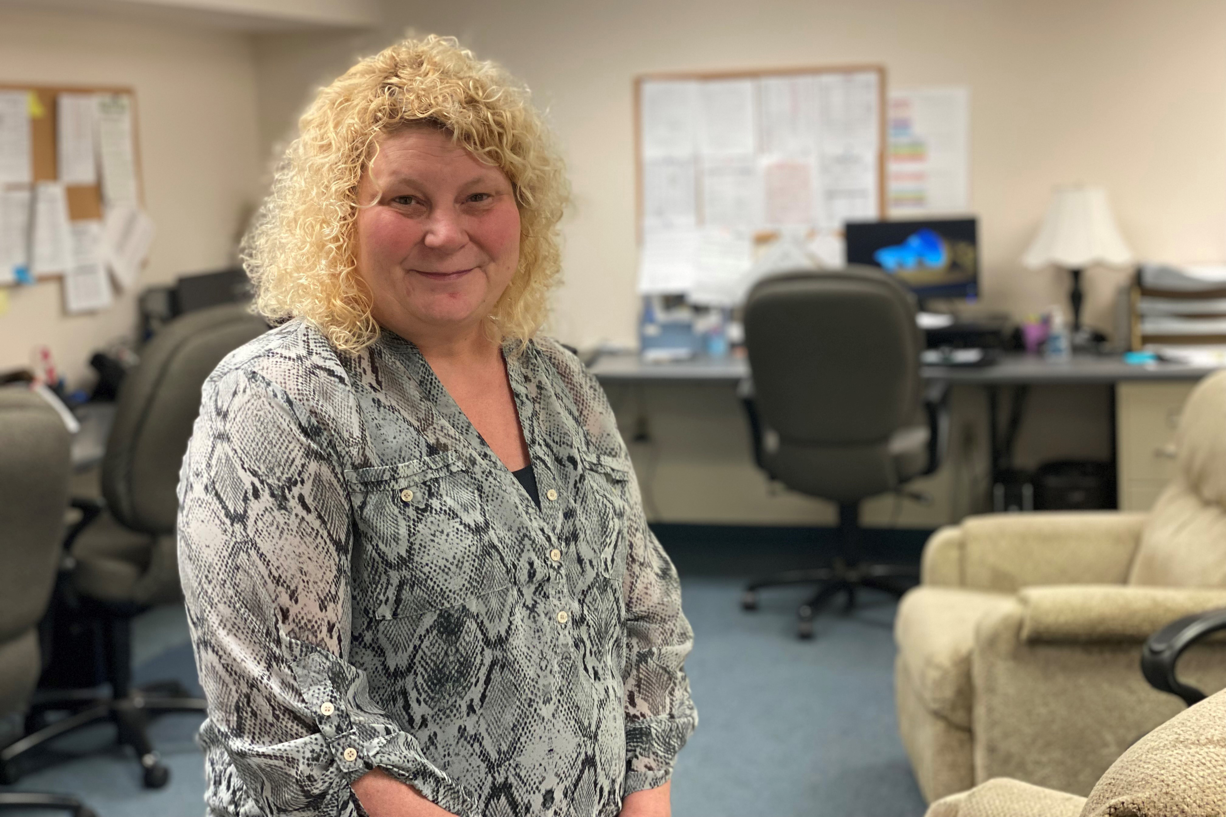 Jayne Wildasin, an adult woman with blonde curly hair wearing a patterned blouse, stands inside her office. A computer desk is visible behind her.