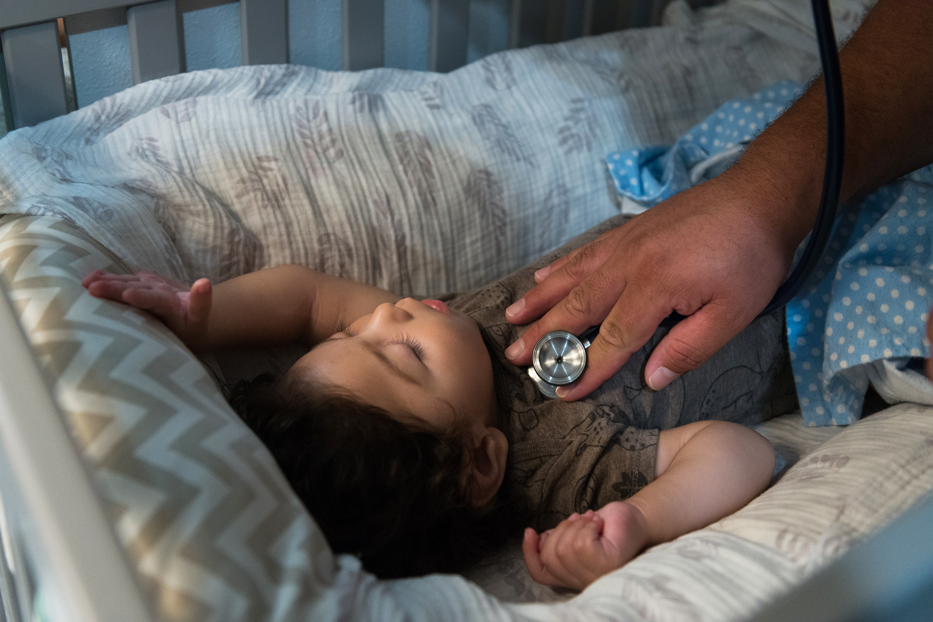 A photo shows a toddler lying asleep in bed, being checked with a stethoscope.