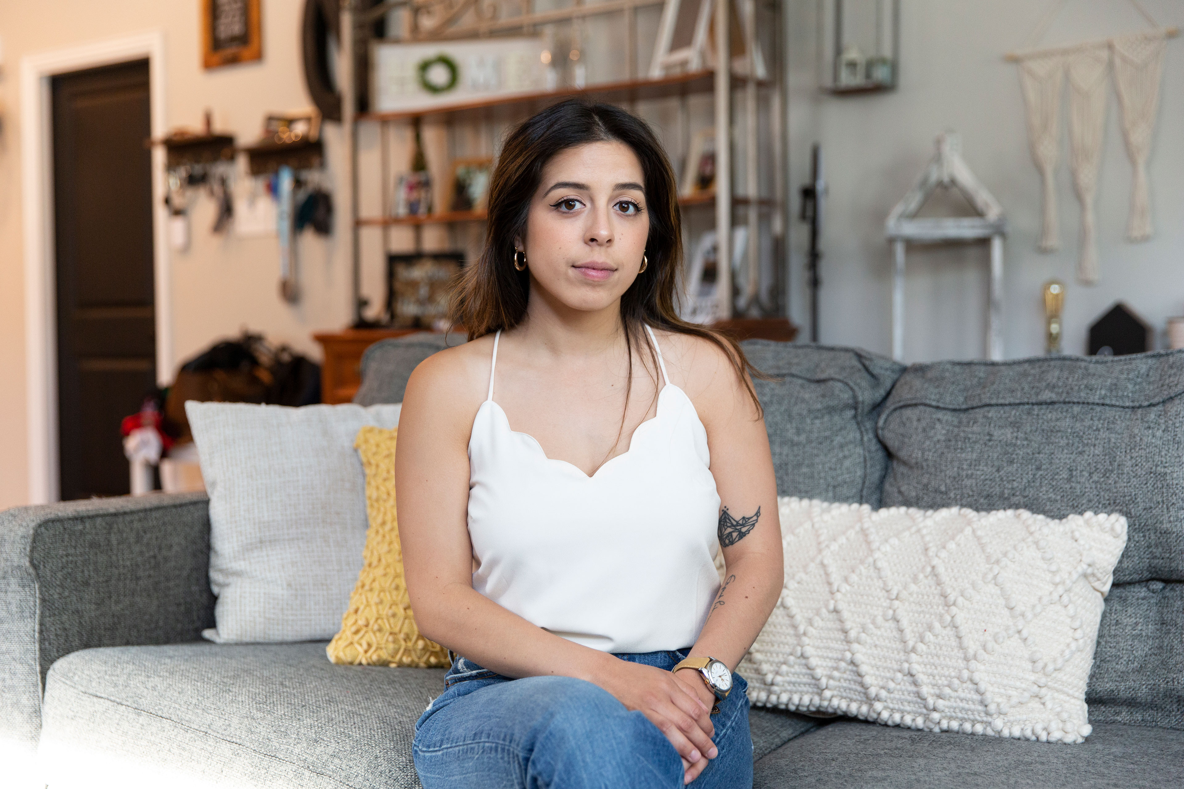 A woman wearing a white tank top and jeans sits on a gray couch in her living room.