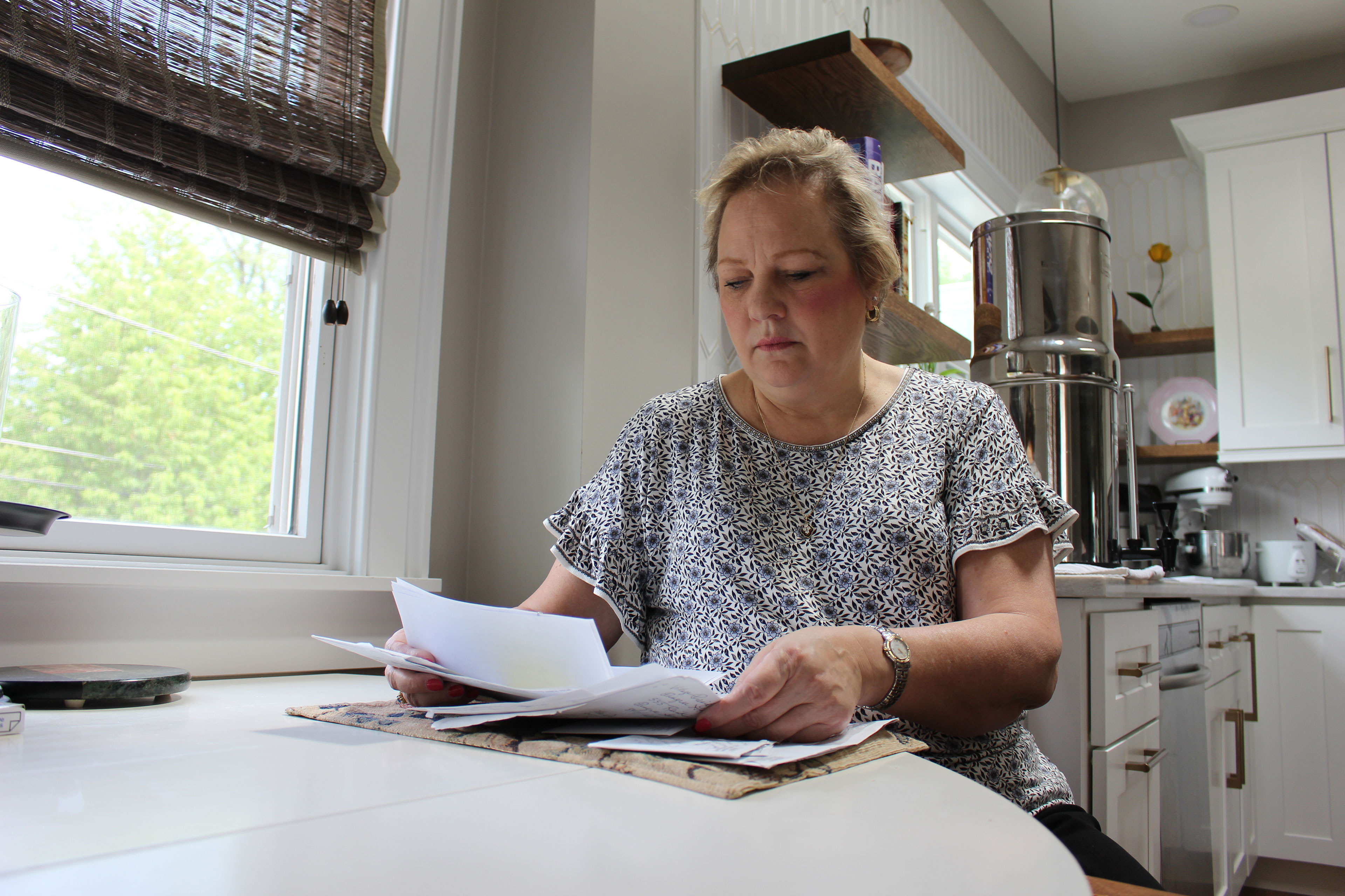 A woman sits at a table next to a window and reads pieces of paper.