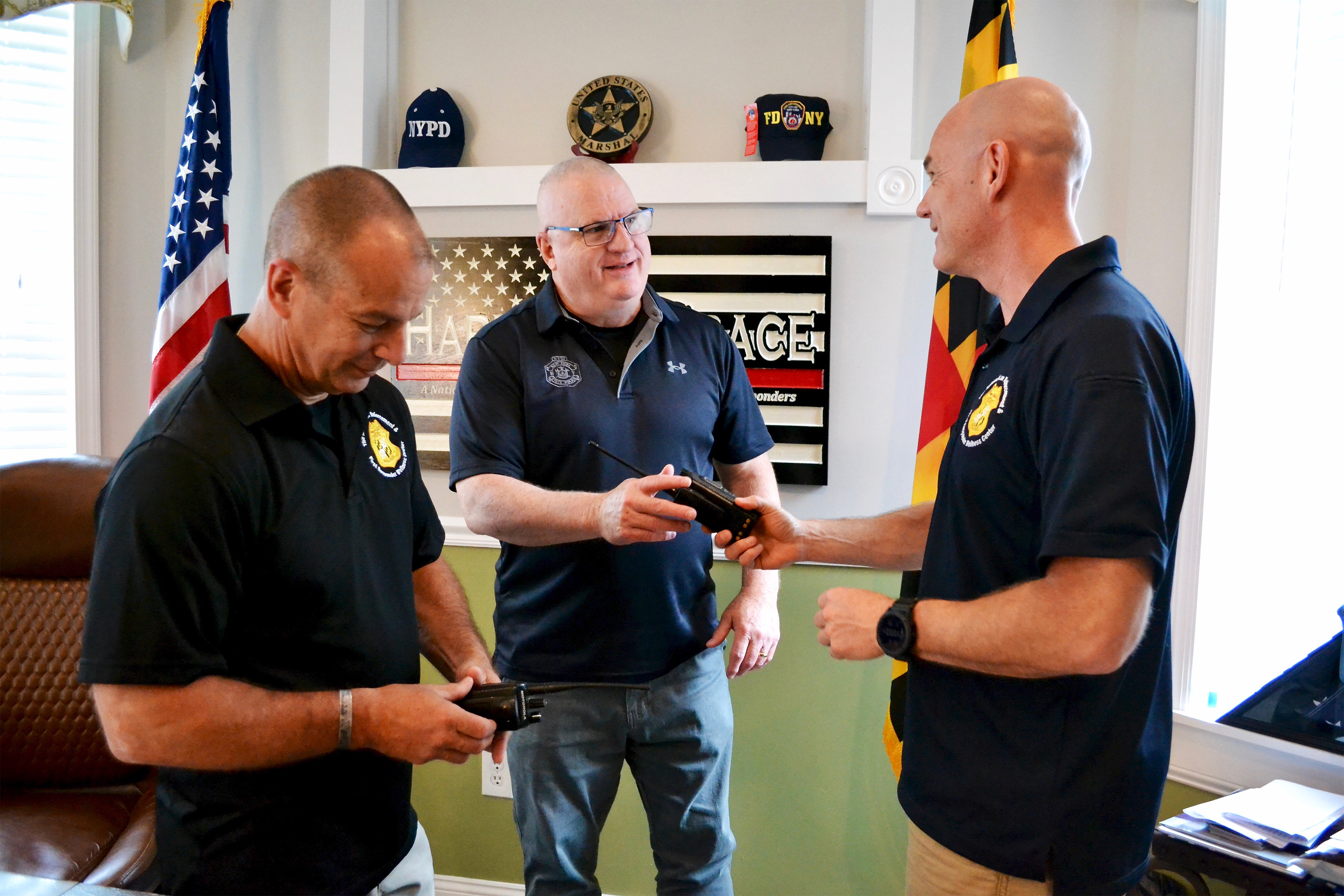 A photo shows three men conversing in a room decorated with law enforcement memorabilia. An American flag and a Maryland flag stand behind them.