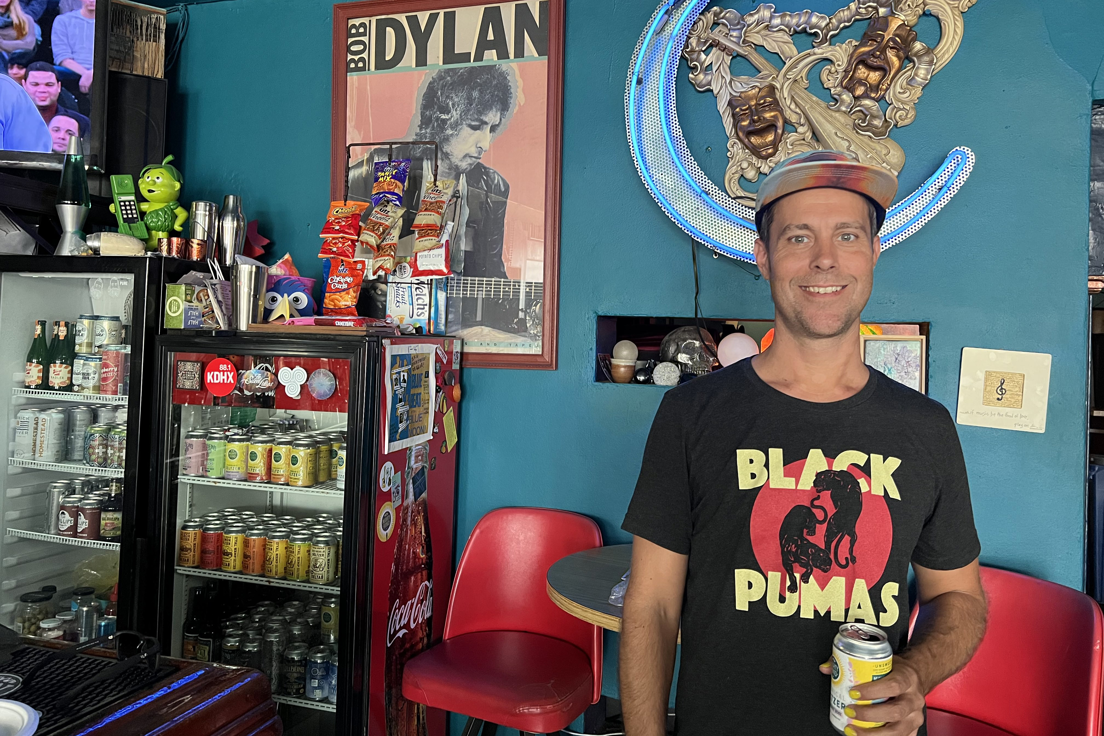 Joshua Grigaitis stands in his shop while holding a can of seltzer and smiling broadly. There are two mini-fridges visible behind him, each filled with seltzer and covered in stickers. A bright red chair and table are below a framed poster of Bob Dylan playing guitar. The walls are painted teal.