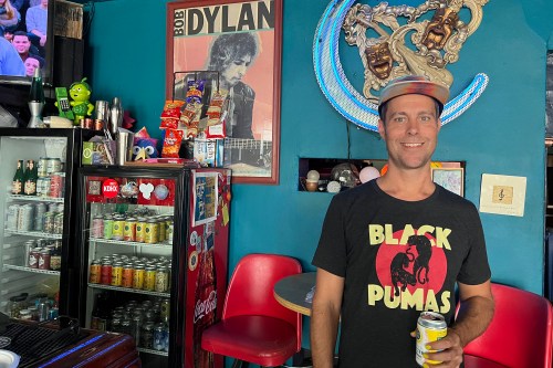 Joshua Grigaitis stands in his shop while holding a can of seltzer and smiling broadly. There are two mini-fridges visible behind him, each filled with seltzer and covered in stickers. A bright red chair and table are below a framed poster of Bob Dylan playing guitar. The walls are painted teal.