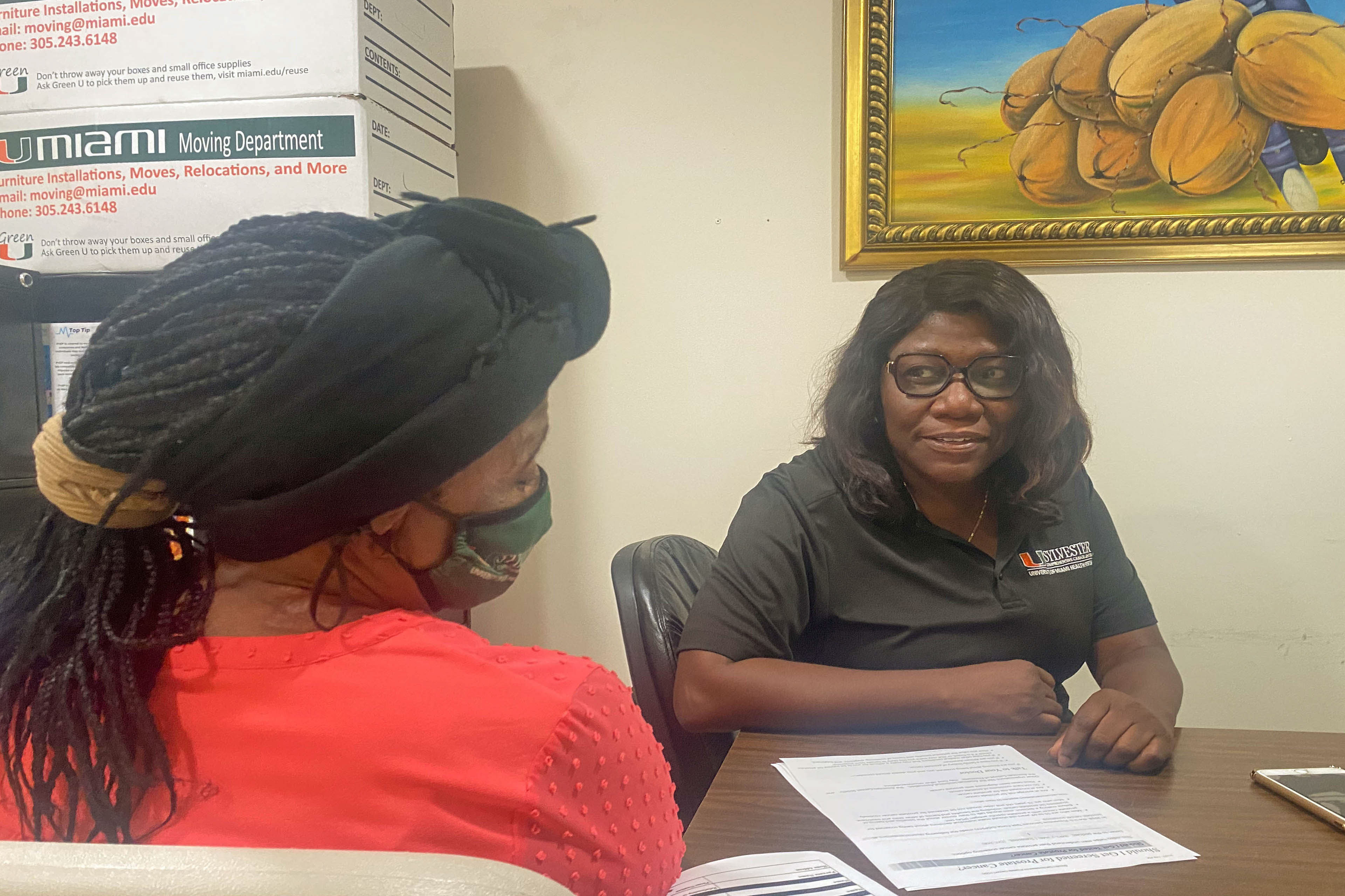 A photo shows Nicole Daceus speaking to Valentine Cesar from across a desk.