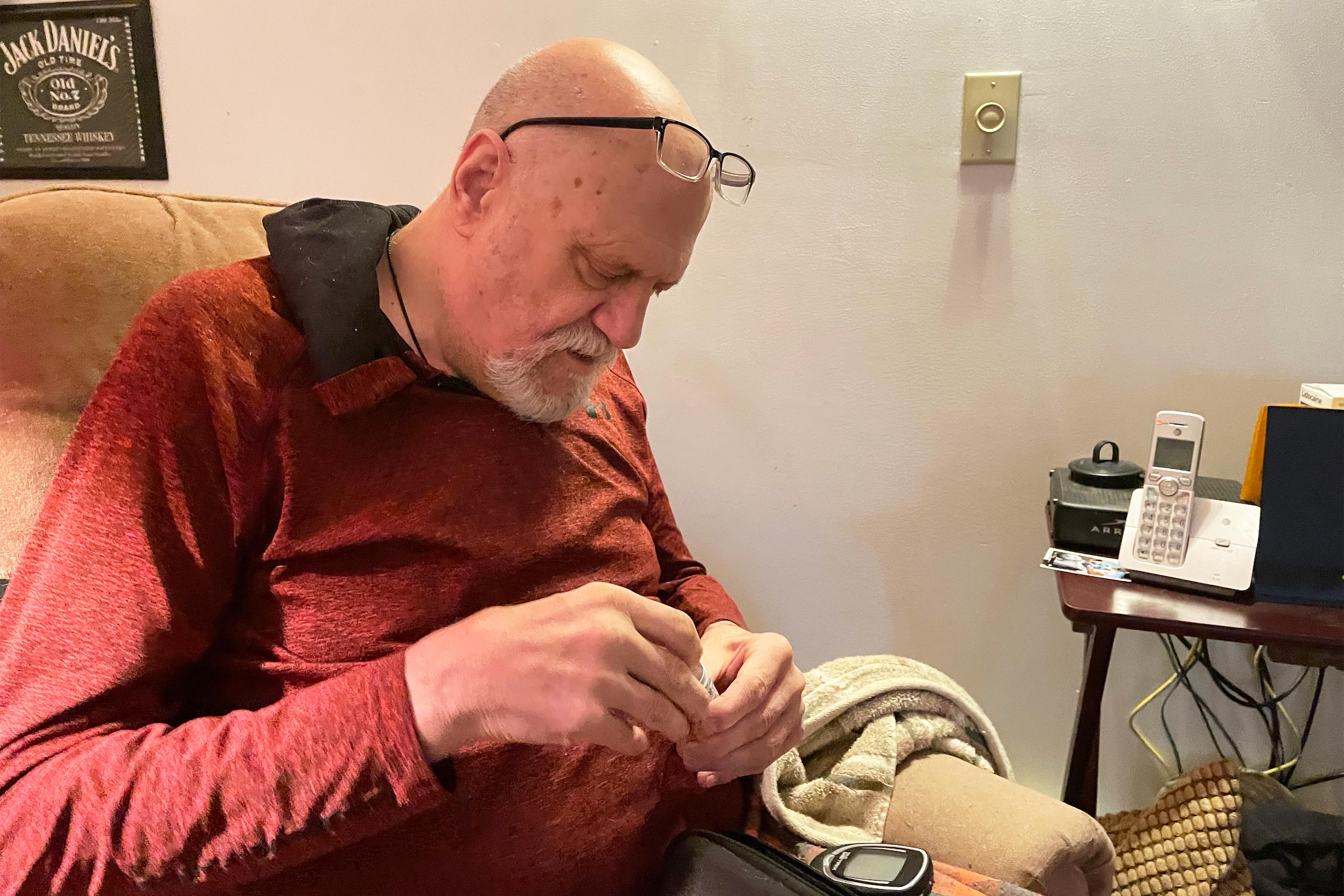 A photo shows Kim Hilton sitting in a chair and checking his blood sugar levels.