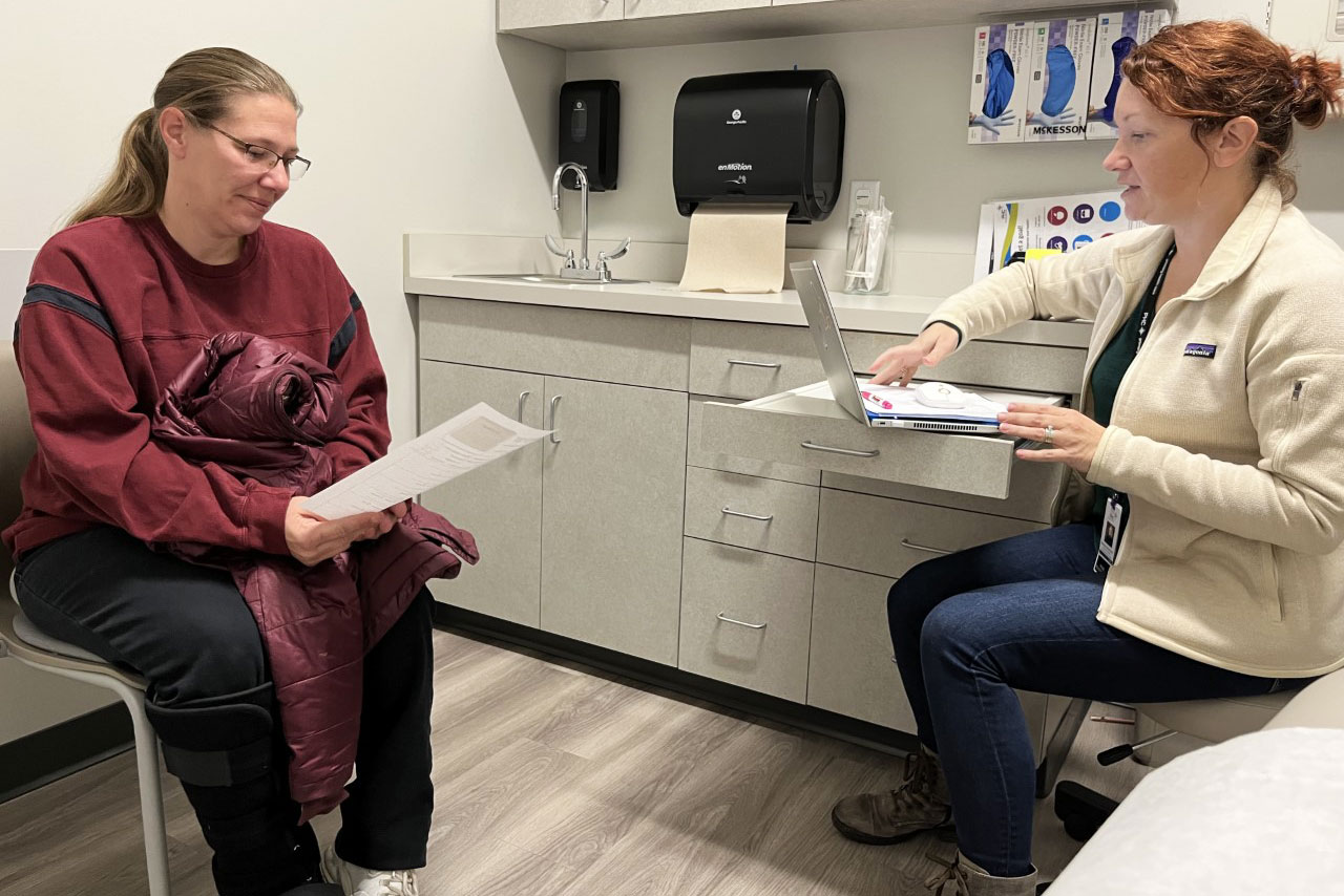 A photo shows Bonnie Purk sitting across from Andrea Storjohann, a nurse practioner, inside a clinic examining room.