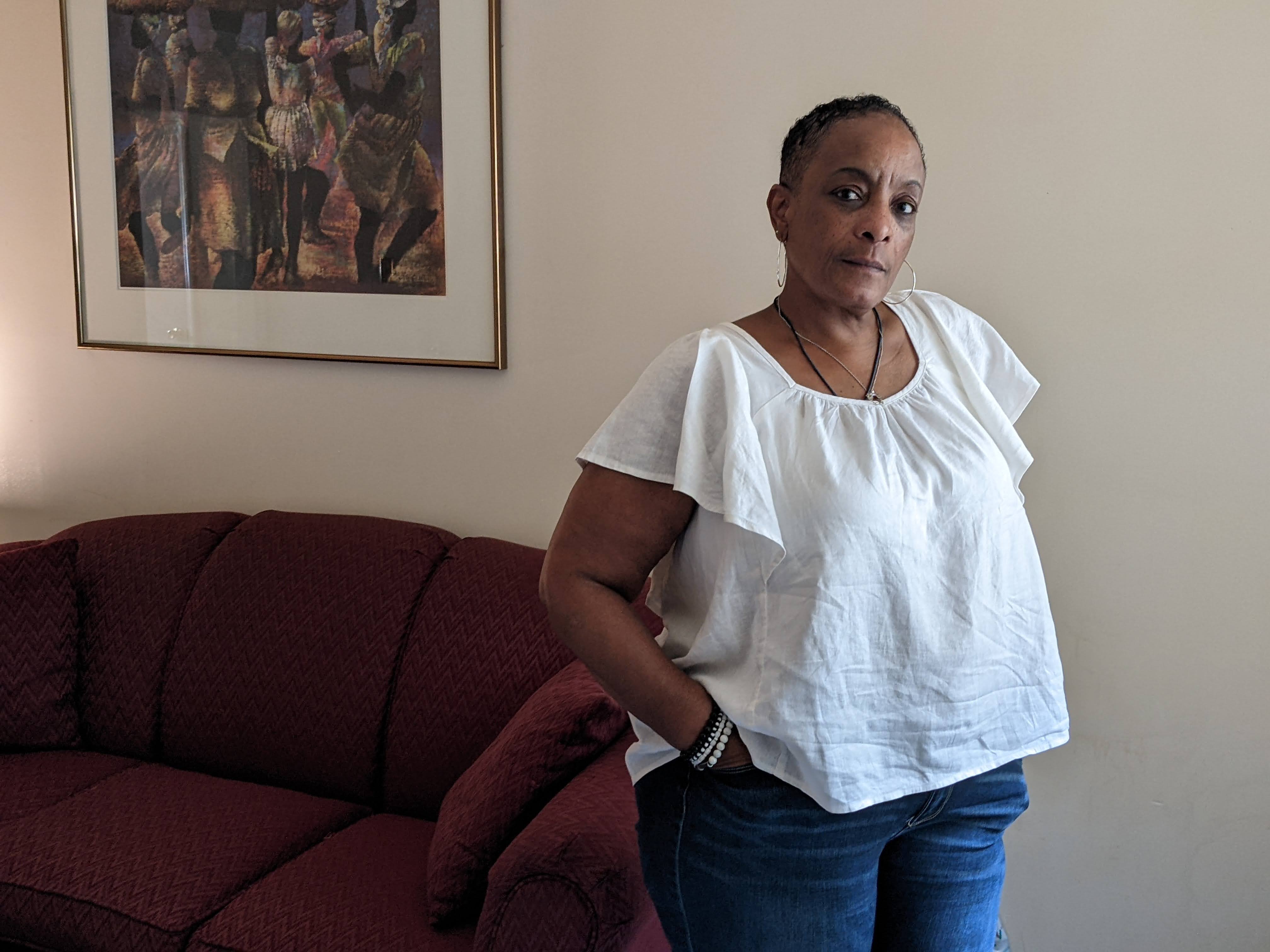 A woman in a short-sleeved white shirt looks at the camer while standing in front of a dark red couch.