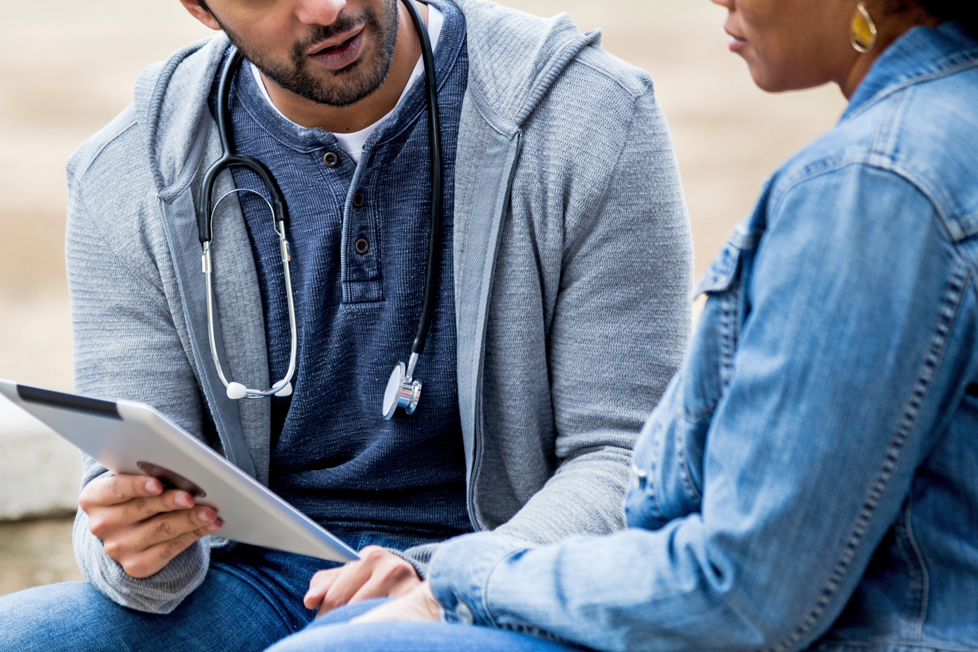 A male medical professional is seen talking to community members outside, showing them information on a tablet screen.