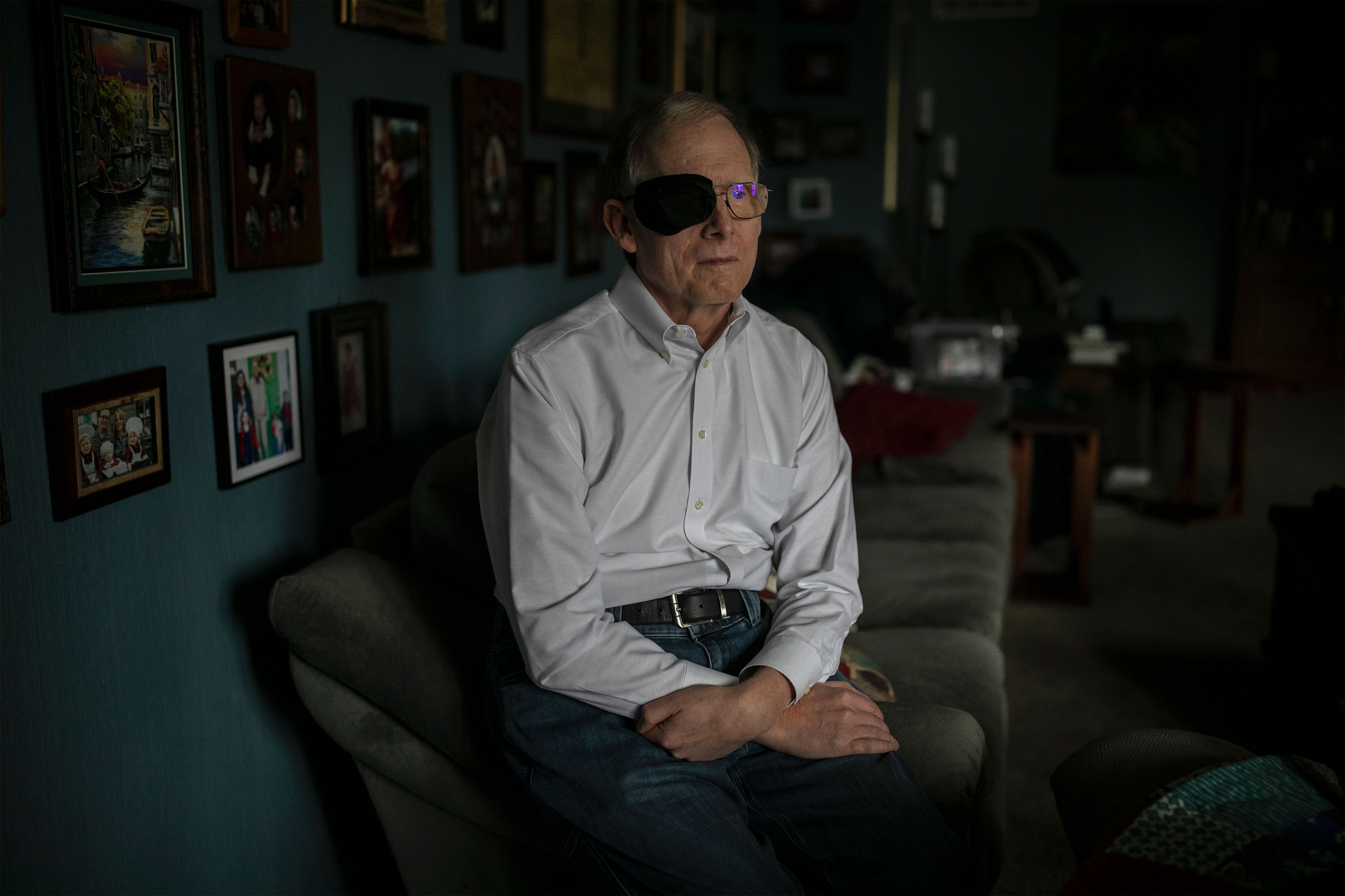 A man with black cloth covering his right eye over his glasses sits on the edge of a couch and looks off-camera to his left.