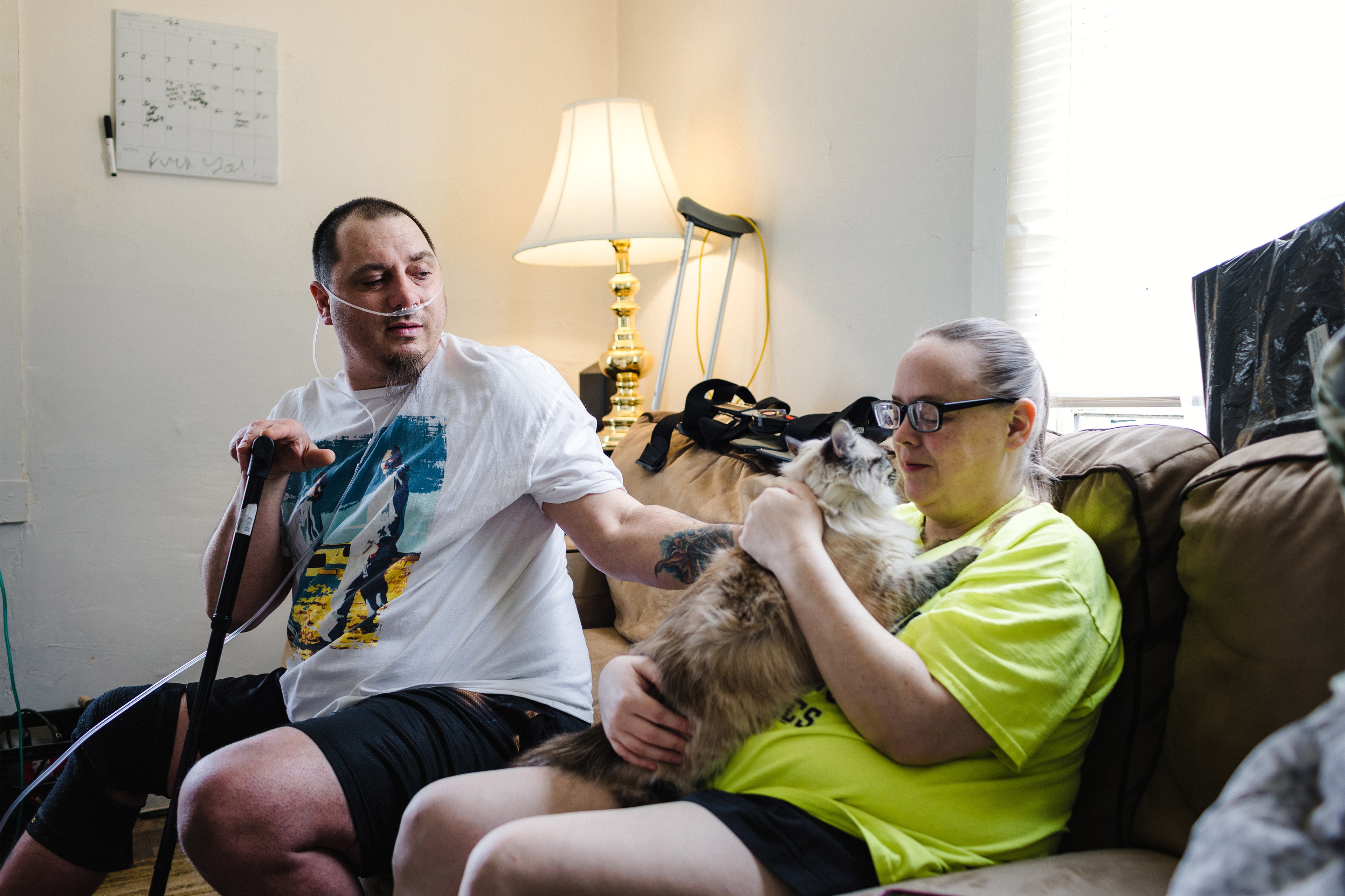A photo shows a man and a woman sitting together on the couch. The two are petting a cat who is sitting in the woman's lap.