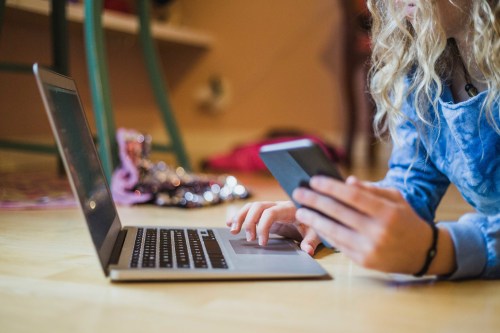 A photograph showing a young girl lying on the floor while using a laptop and smart phone at the same time.