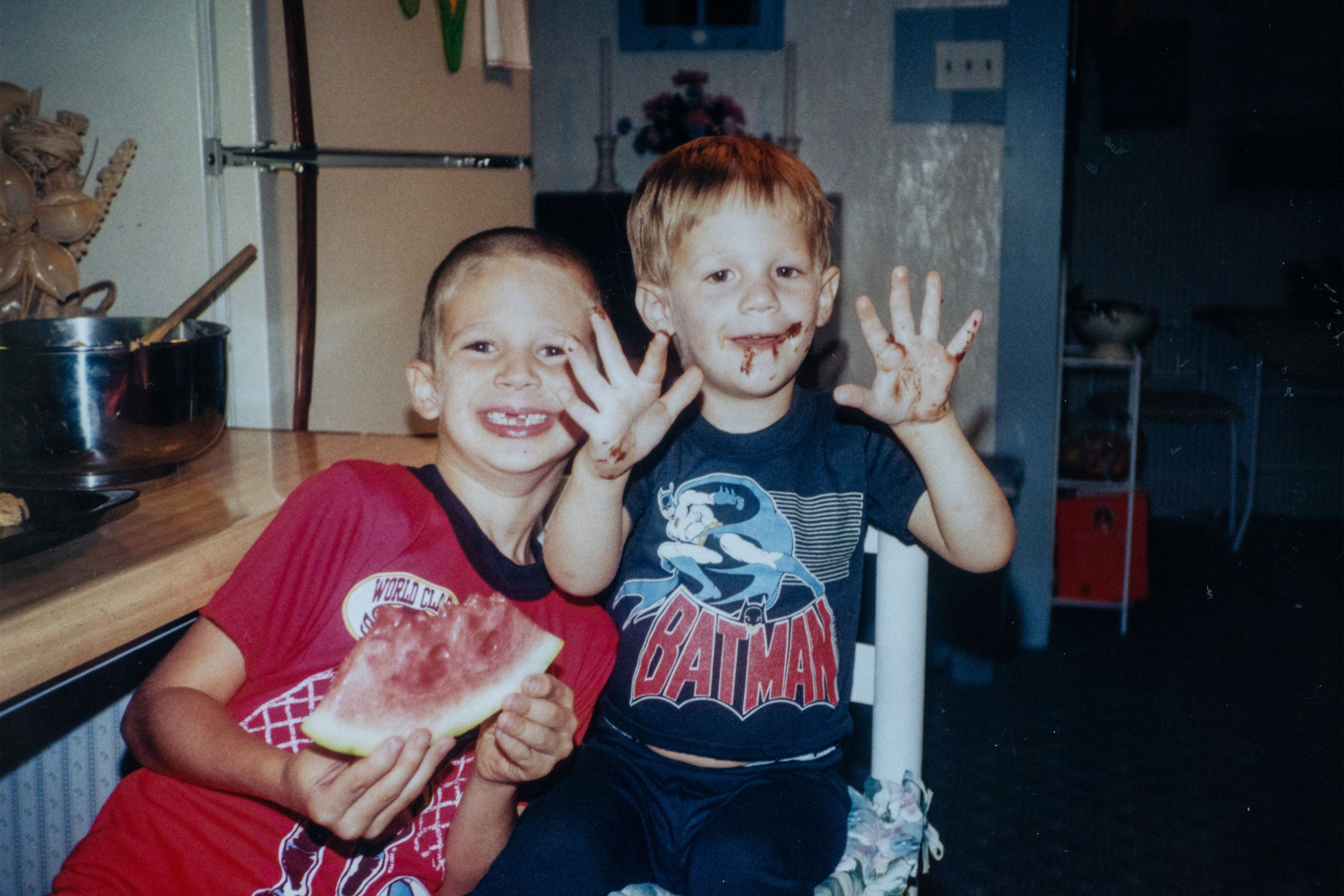 Two children, both boys, smile at the camera. One boy, in a red tshirt, is eating a slice of watermelon and has several front teeth missing. The other, in a navy Batman t-shirt, holds up his hands which, like his face, are covered in food.