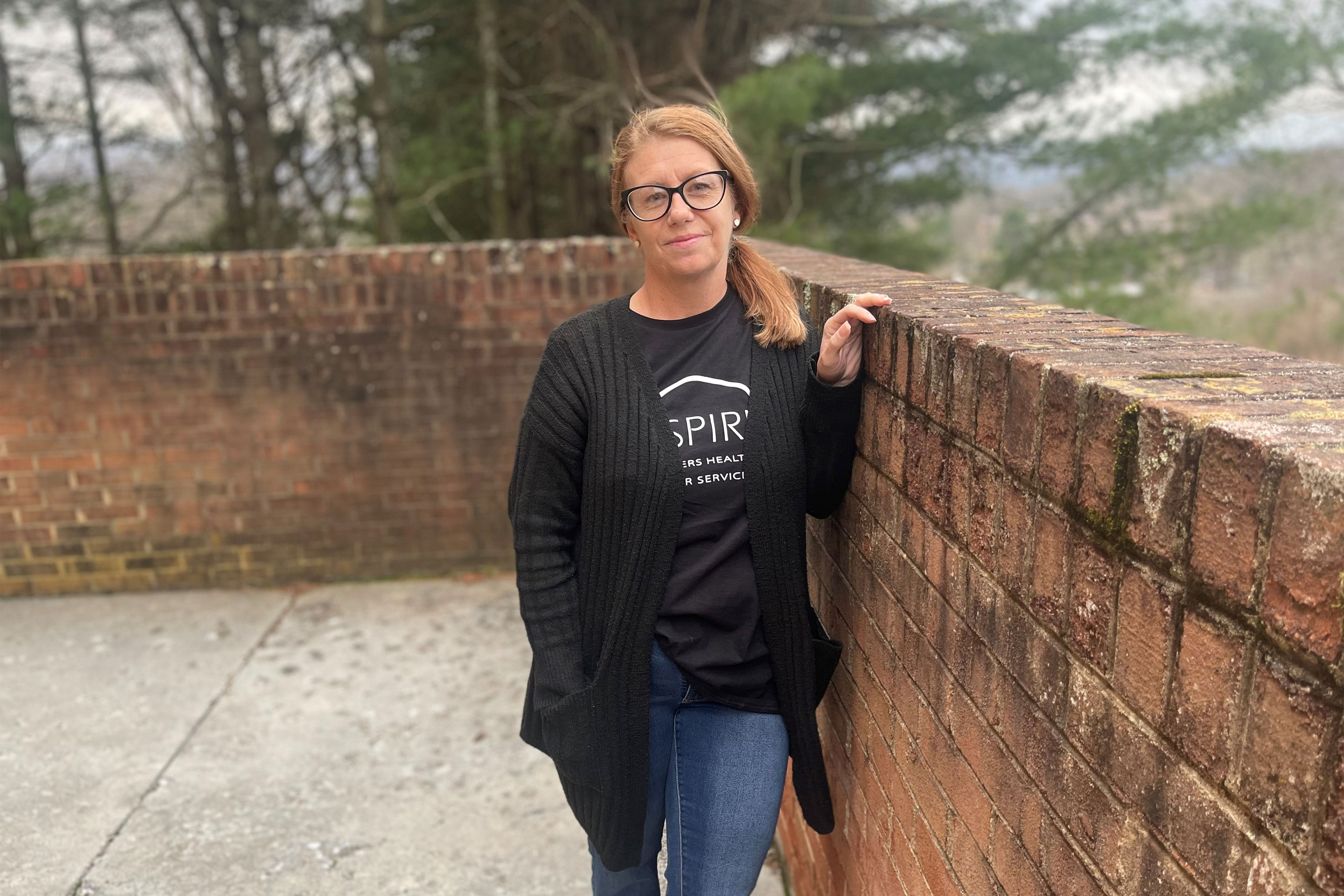 A woman with light hair and black cat-eye shaped glasses leans against a brick wall outside. She is looking directly at the camera with a slight smile on her face and is wearing a black t-shirt with white writing, jeans, and a black cardigan.