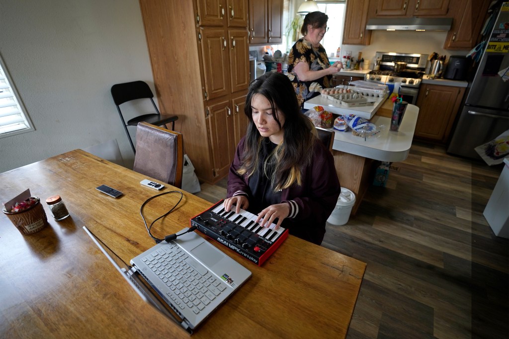 A photo of teenager using her keyboard and laptop to make music while her mother cooks in the kitchen behind her.