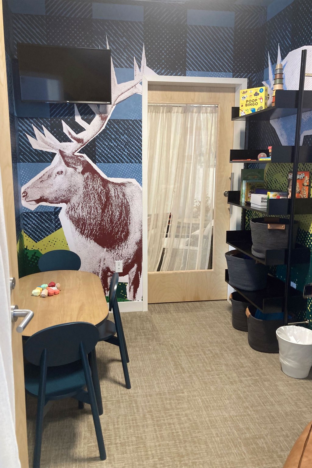 There are child-sized table and chairs and shelves with games in this small room. There is a tv mounted on the wall and an large image of an elk is affixed to the the wall.