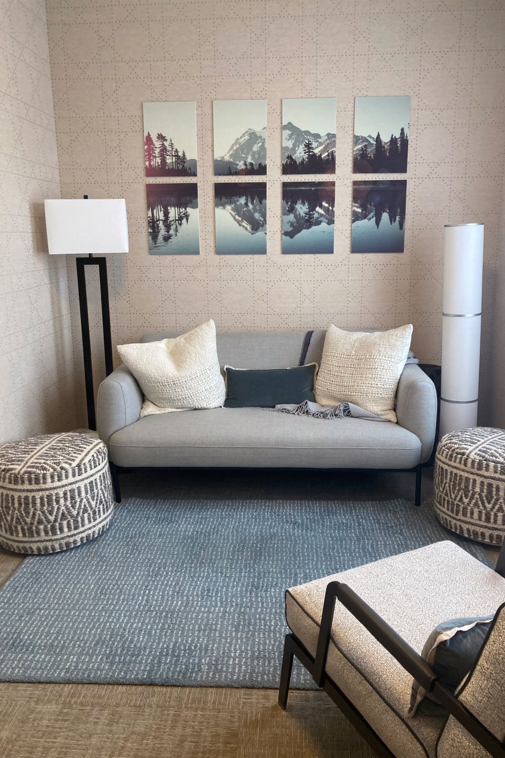 A room furnished with a sofa, ottomans, a chair, and lamps, has 8 panels decorating the wall which together show a mountain scene reflected in a lake.