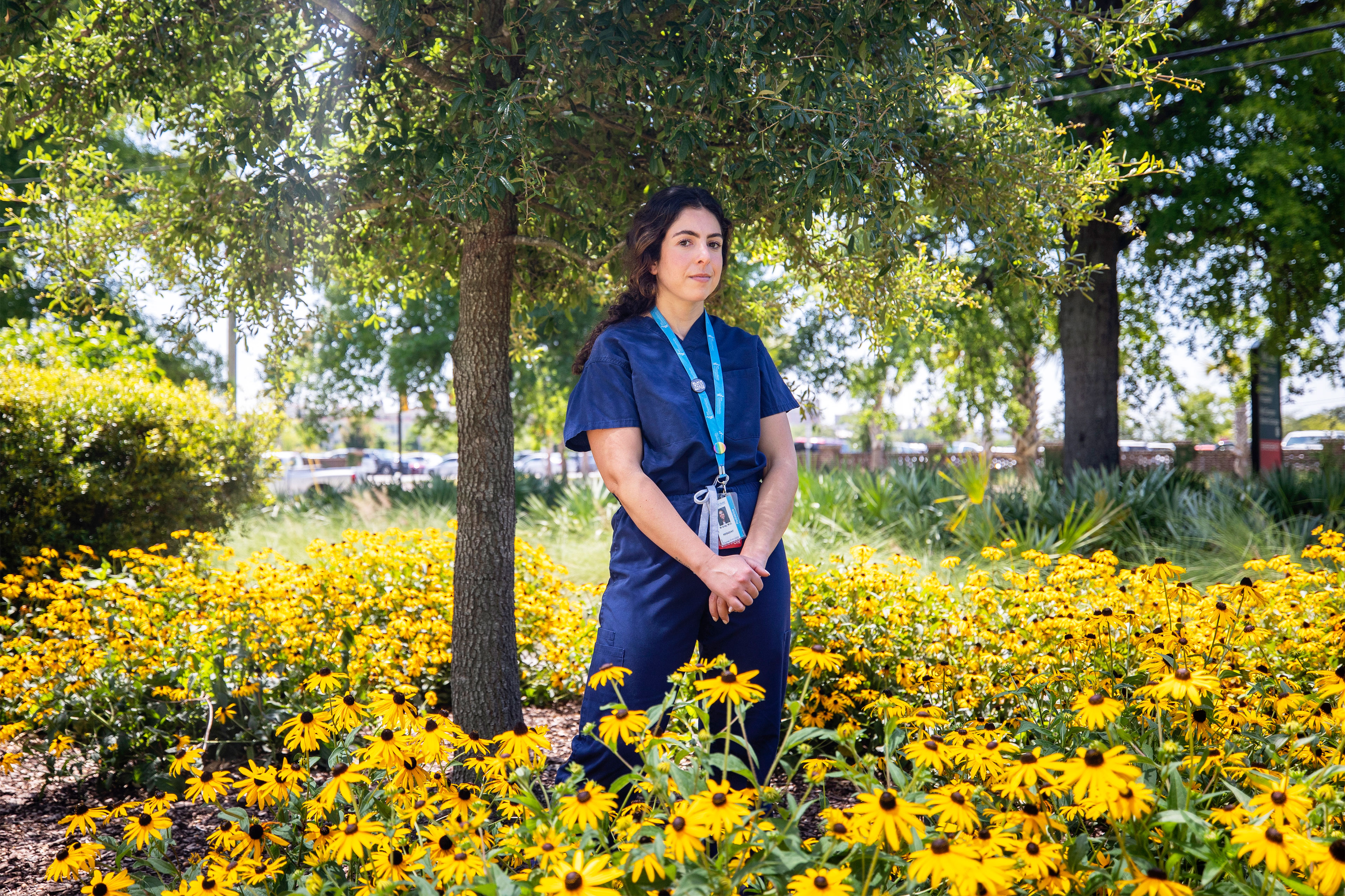A photo of Melanie Gray Miller standing outside by a tree and surrounded by flowers.