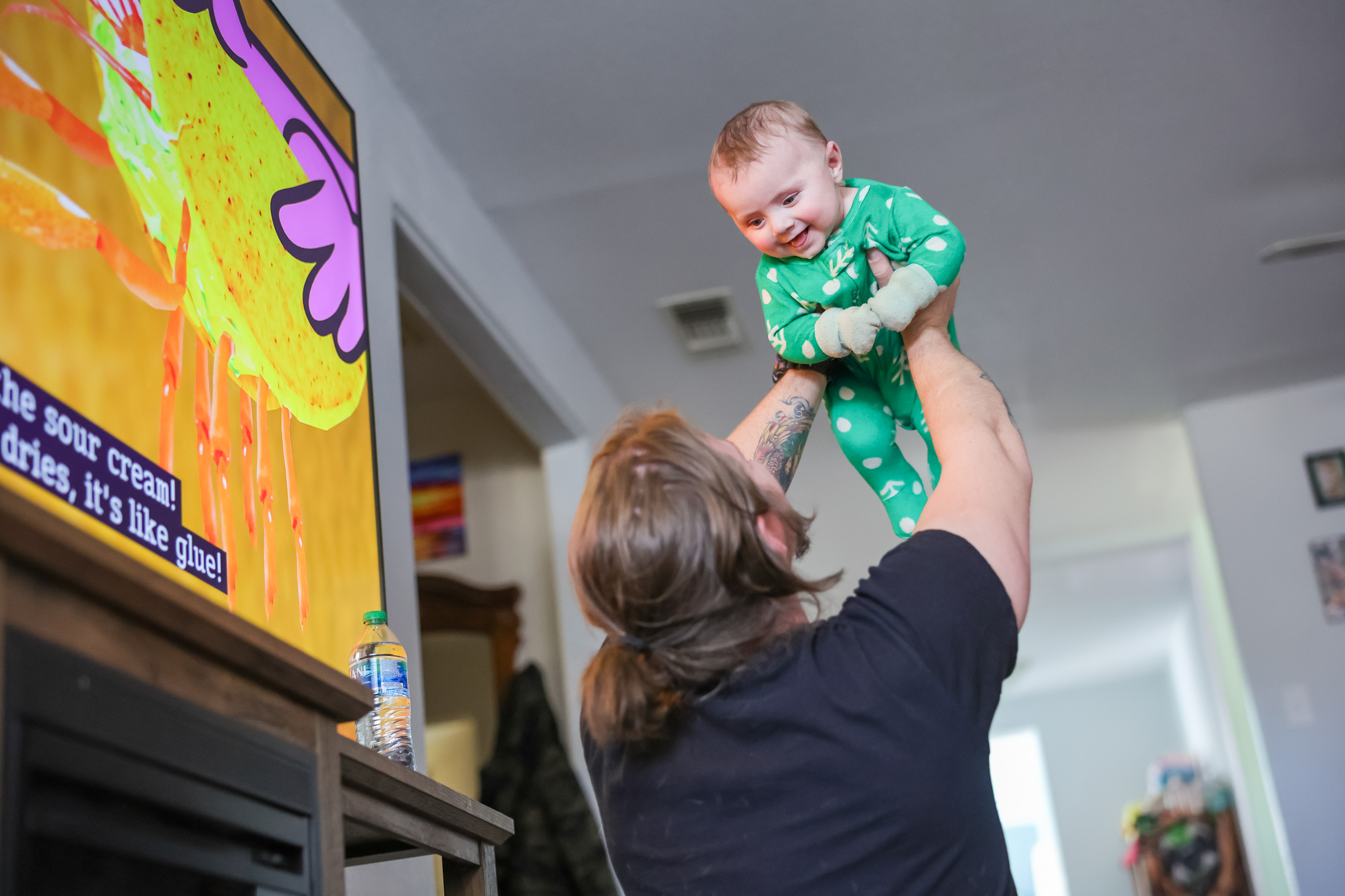 Zane Vandiver holds his 6-month-old son, Ezra, up above his head. Erza is wearing a green onesie and is smiling down at his father.
