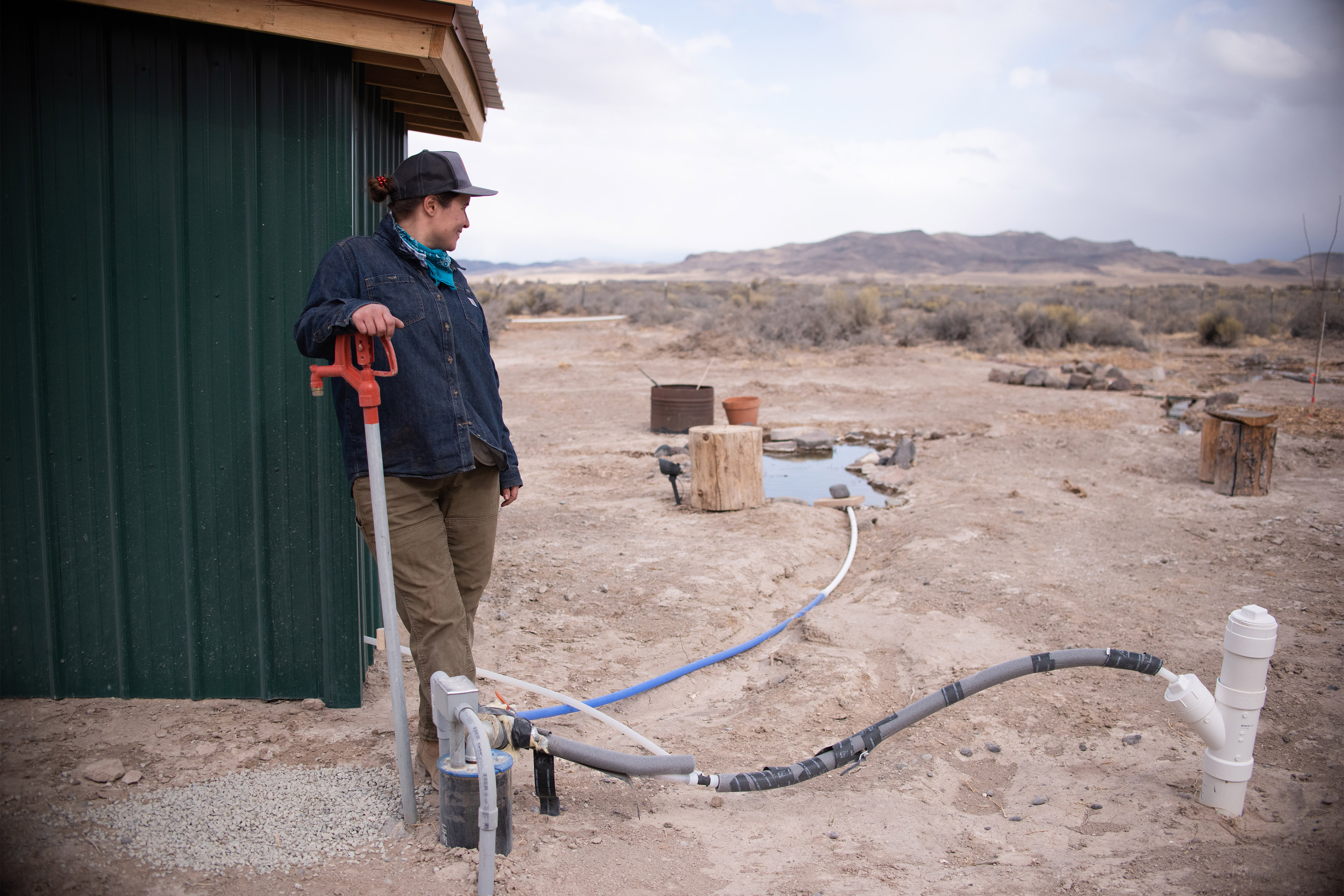 A photo of a woman outside leaning against a shed and looking at her drinking well.
