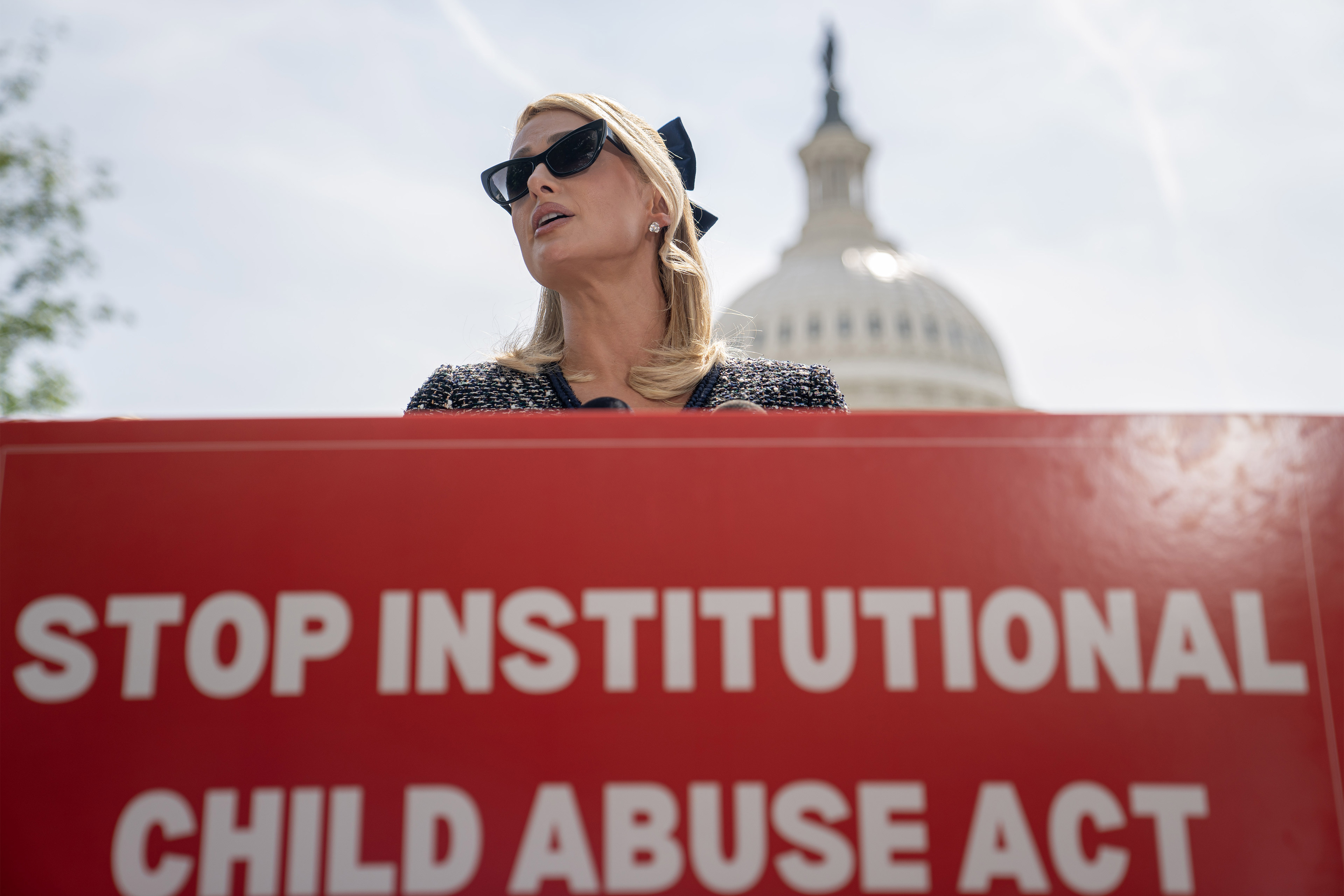 A photo of Paris Hilton standing behind a sign that reads, "Stop Institutional Child Abuse Act." The U.S. Capitol dome is seen behind her.