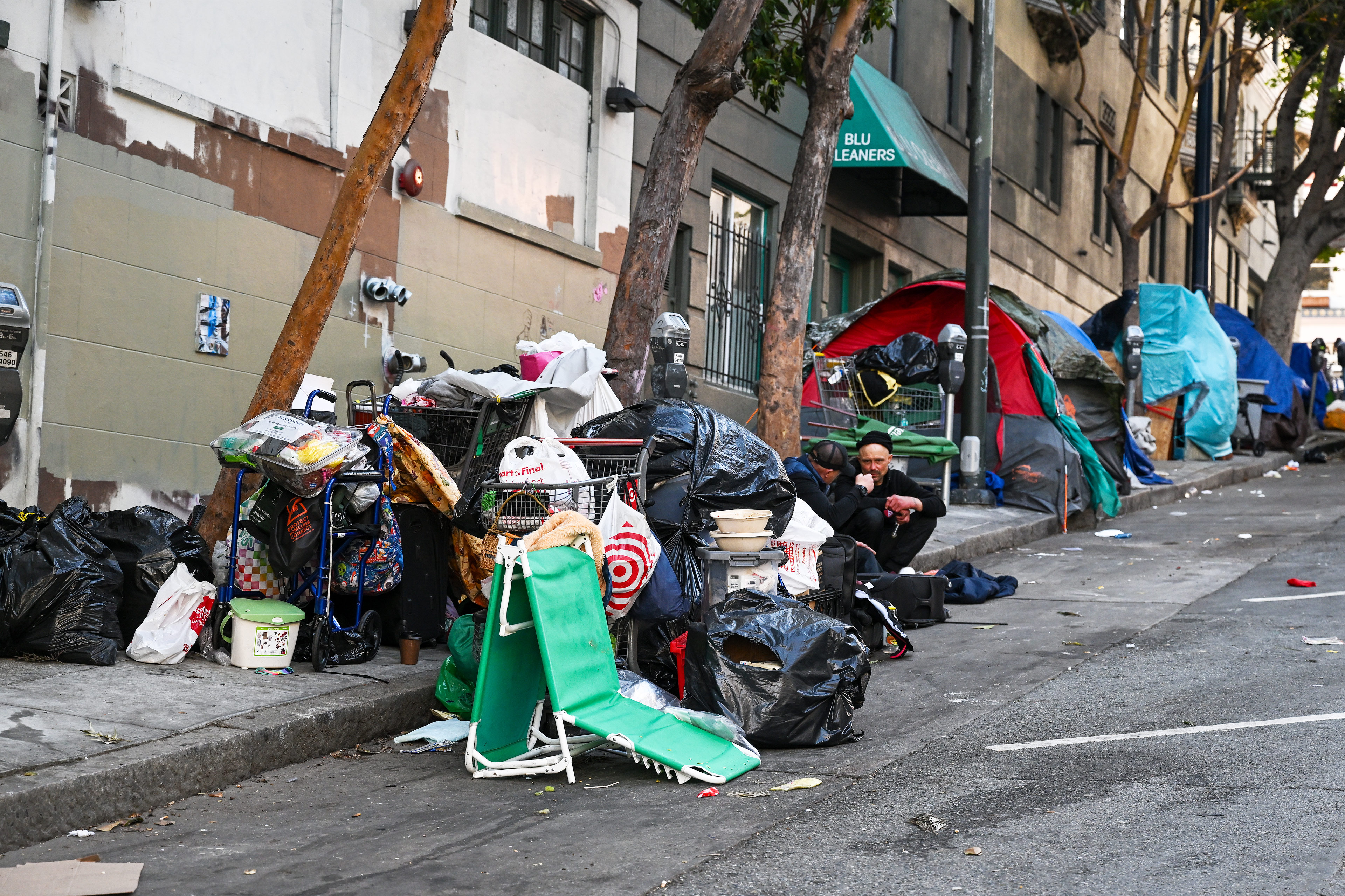 A photo of two people sitting on the sidewalk surrounded by trash bags and grocery carts filled with supplies.