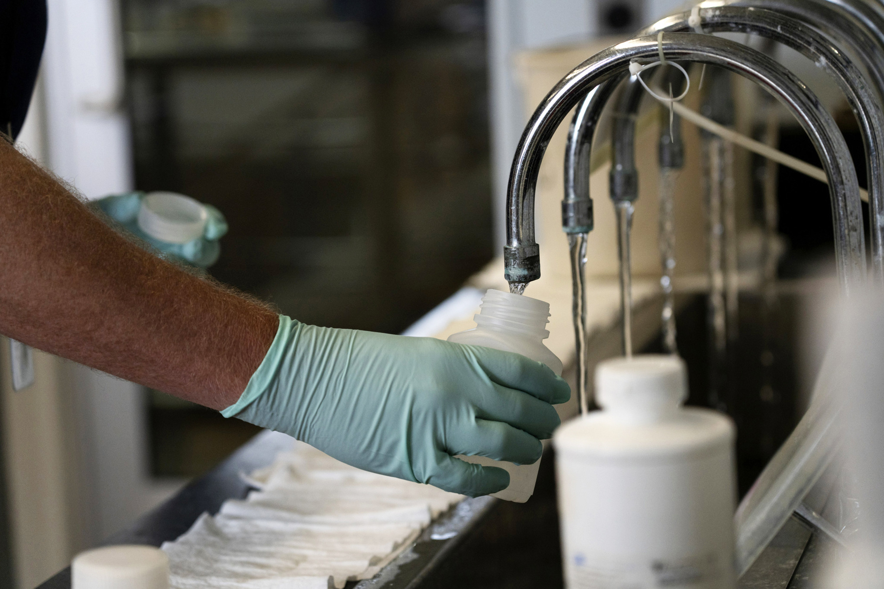 A photo of a gloved hand collecting a sample of water from a faucet in a lab.