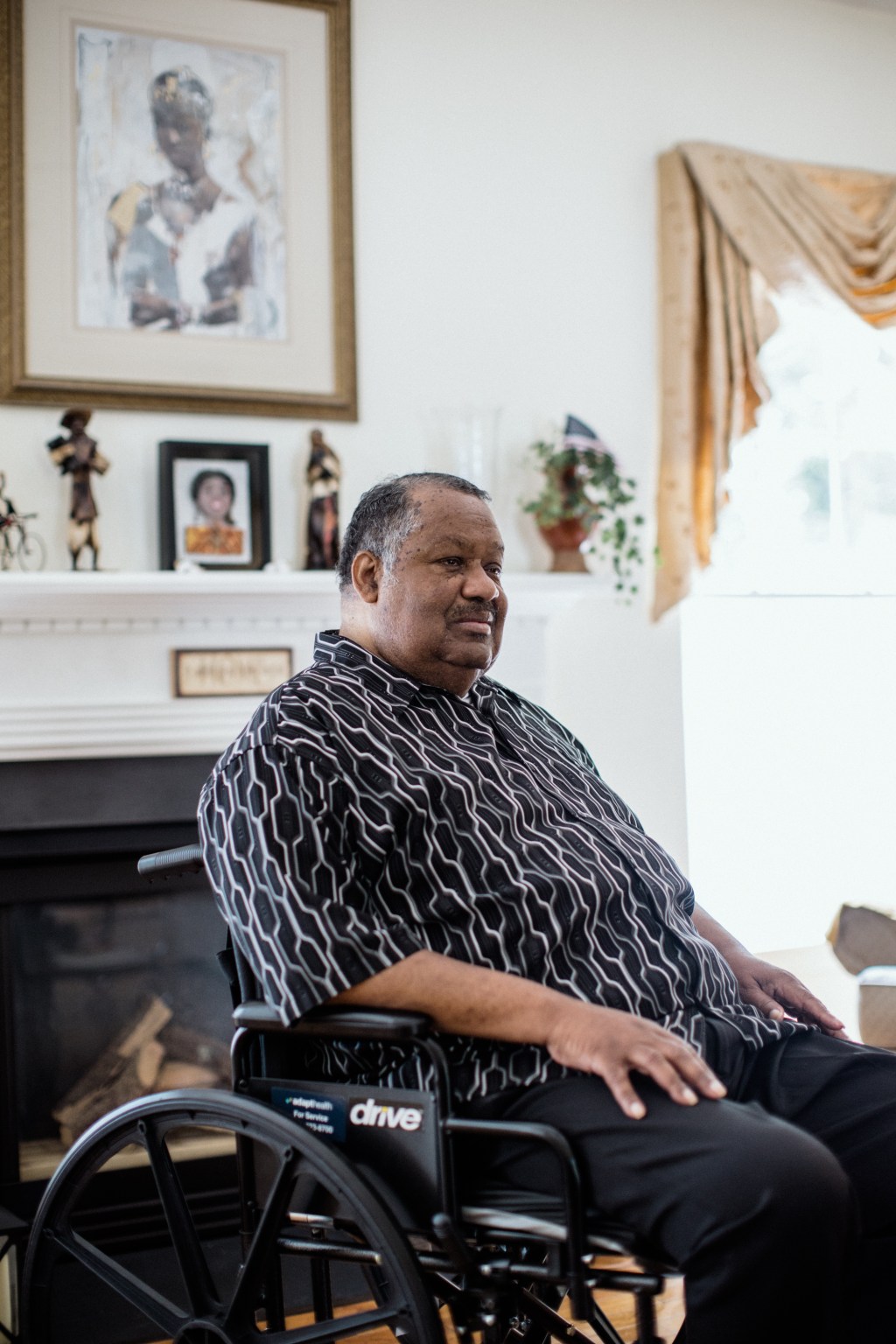 Thomas Greene is sitting in a wheelchair in his home, which is brightly lit by sunlight. He looks away from the camera.