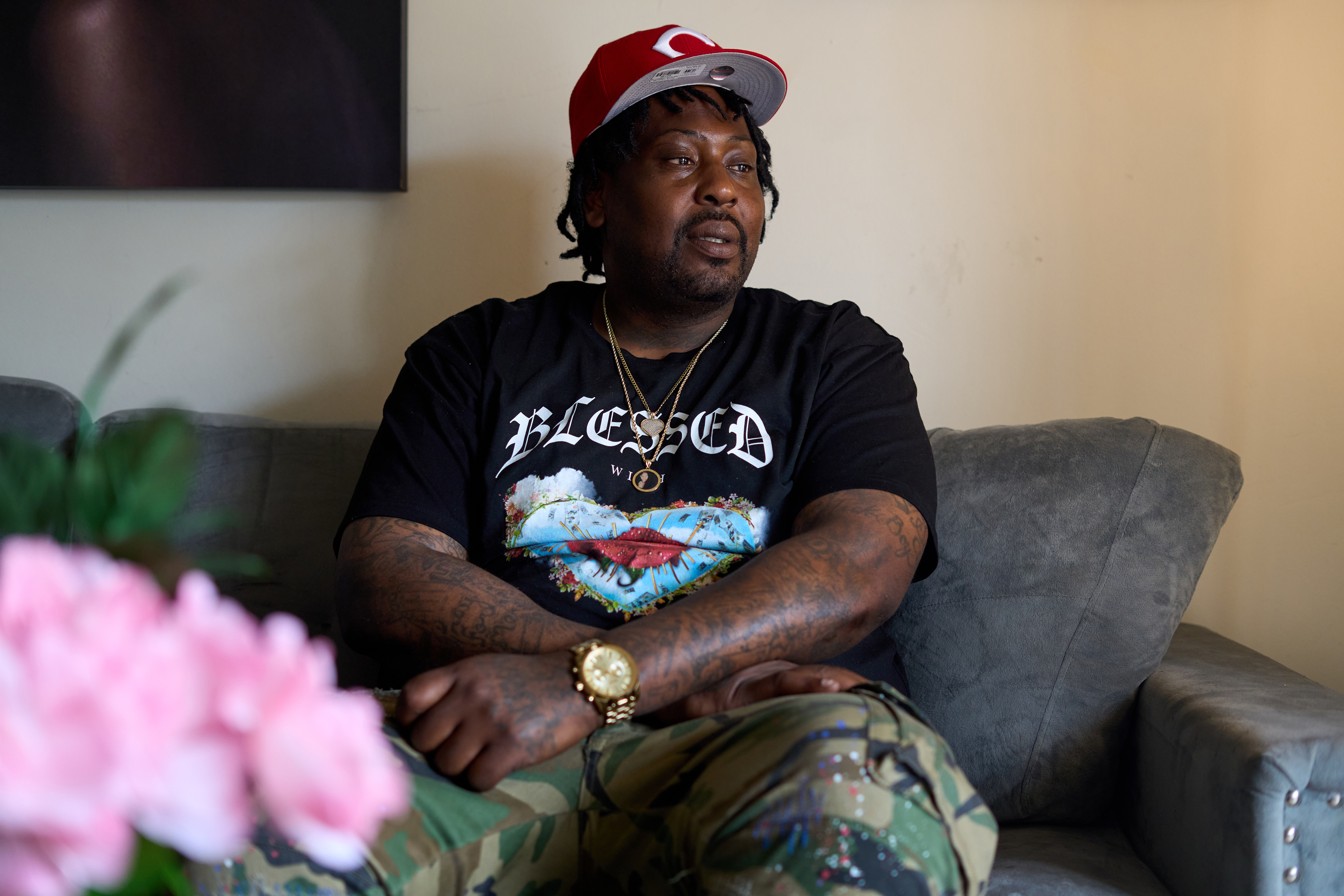 A man in a black t-shirt with the word 'Blessed' across the chest in white, looks to the left with a sombre expression on his face. His arms are tattooed and he has a gold watch around his wrist. 