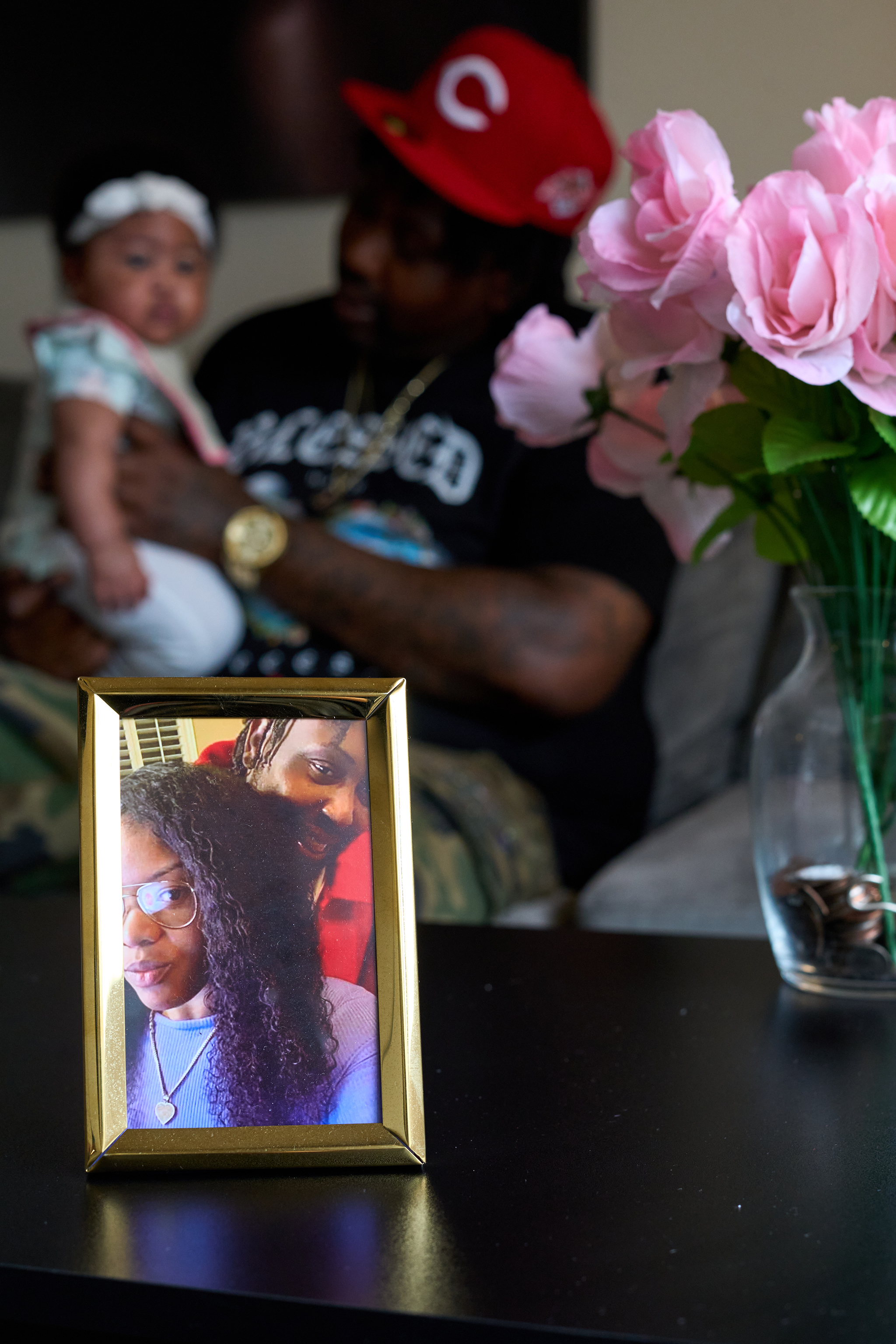 In the foreground is a vase of flowers and a gold framed photos of a man and woman on table. In the background, out of focus, is a father holding his baby daughter on his knee. 