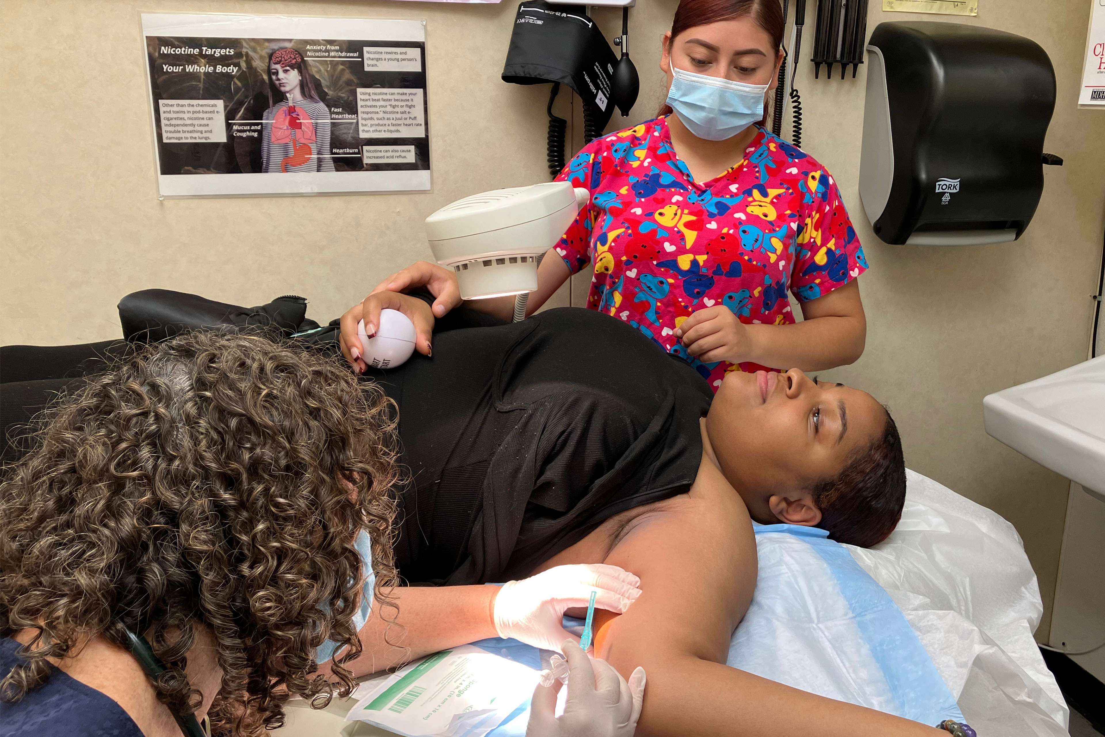 A photo of a young Black woman lying down as two medical workers assist her.
