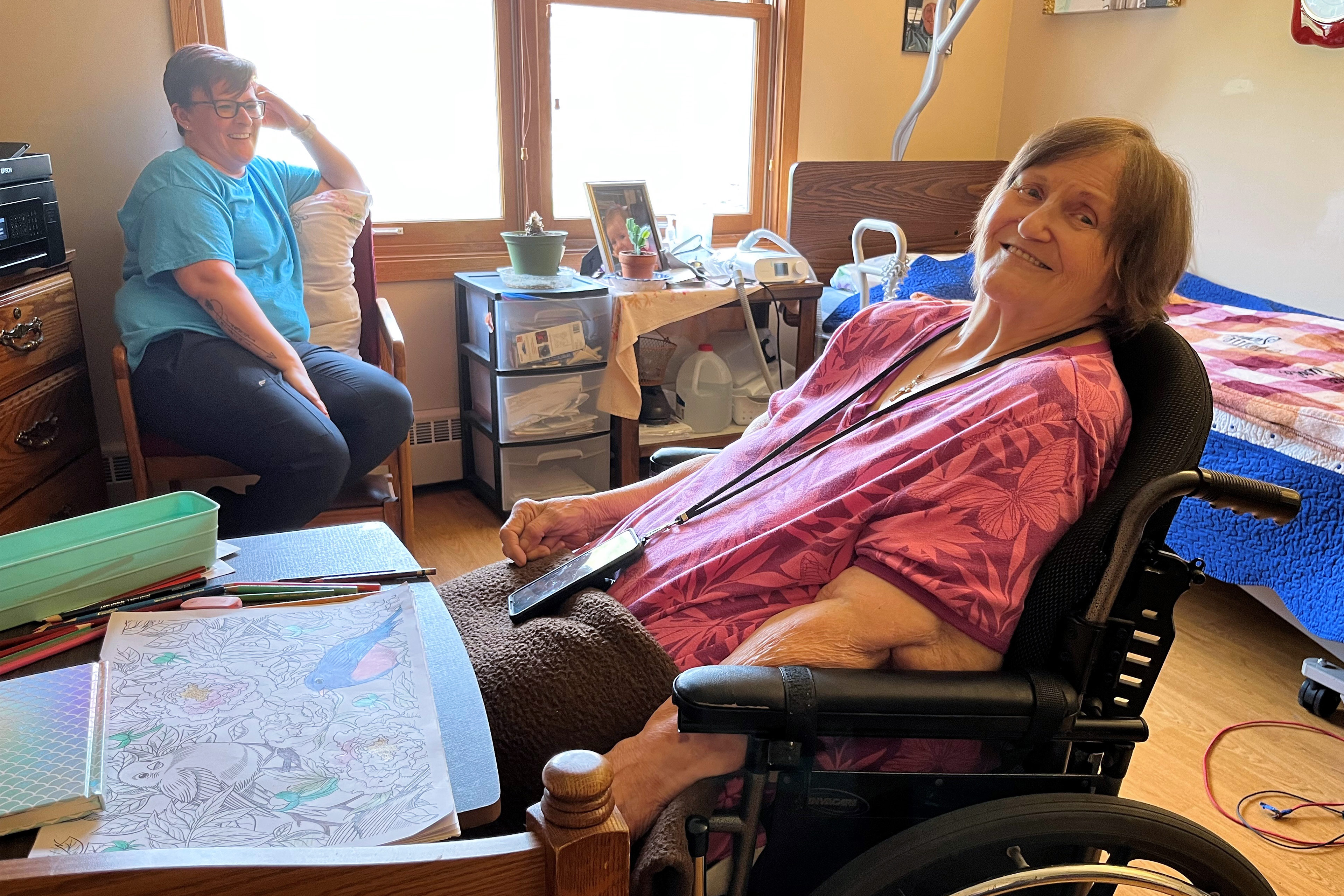 A photo of an older woman sitting at a desk in a wheelchair. Another woman is sitting in the background behind her.