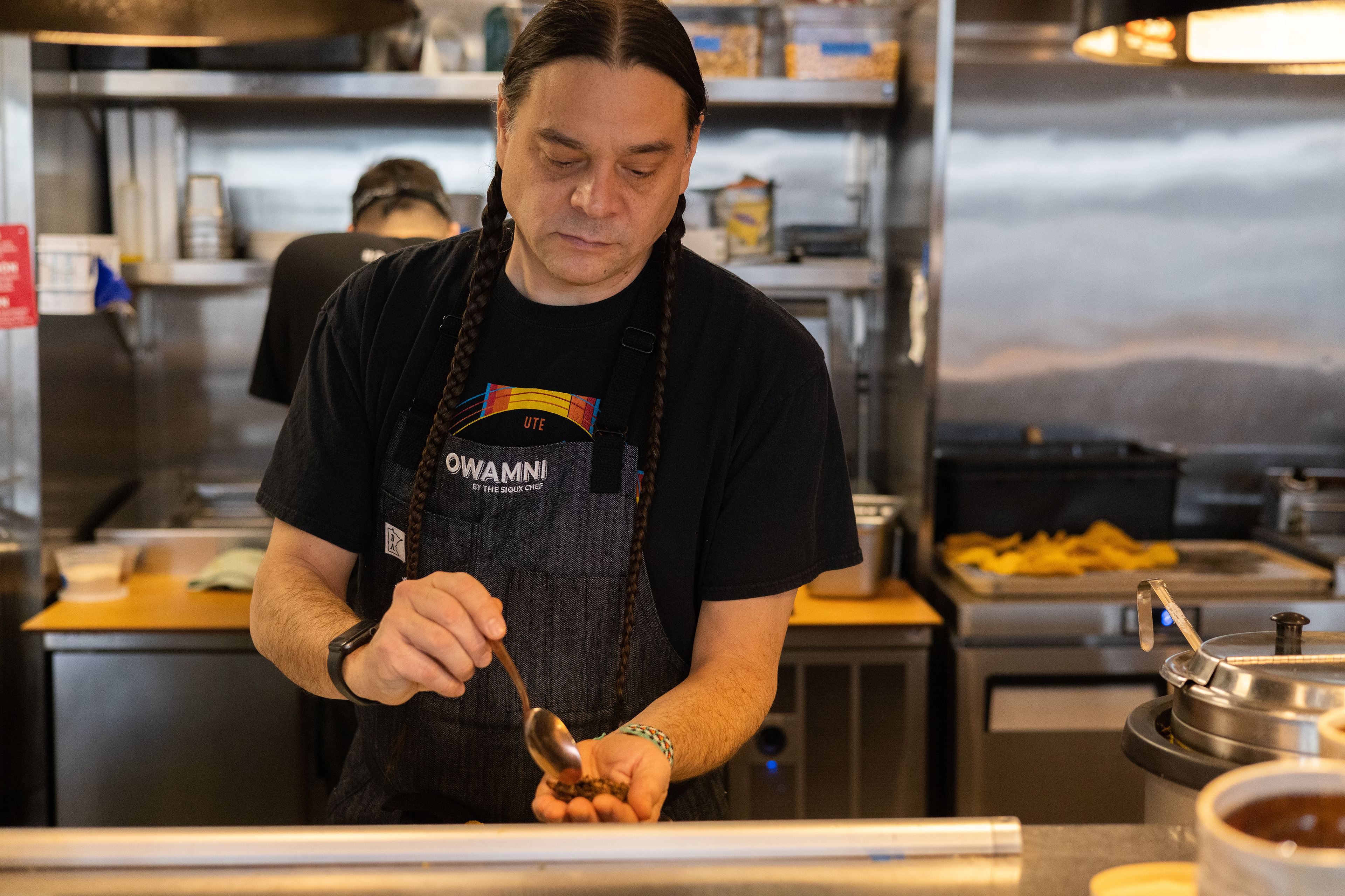 A man holds a spoon in his right hand over his cupped left hand as he stands in a restaurant kitchen wearing an apron
