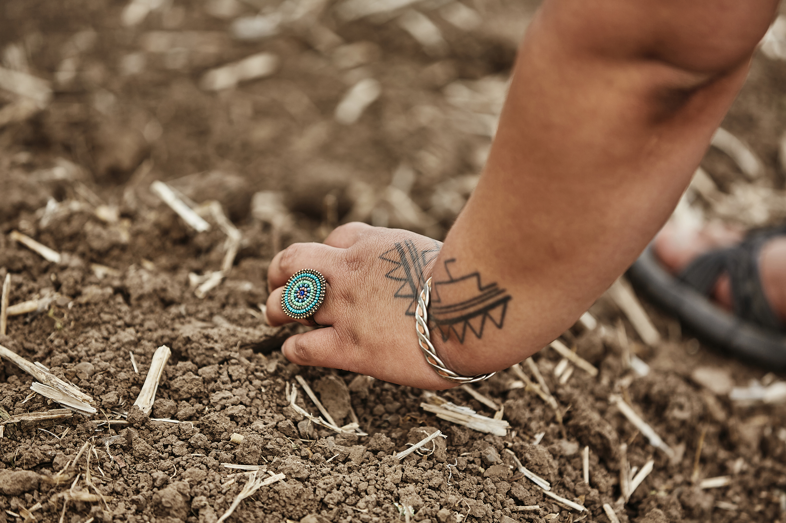 The tattooed hand of a woman, wearing a circular turquoise ring and a bracelet, digs in dirt in a garden.